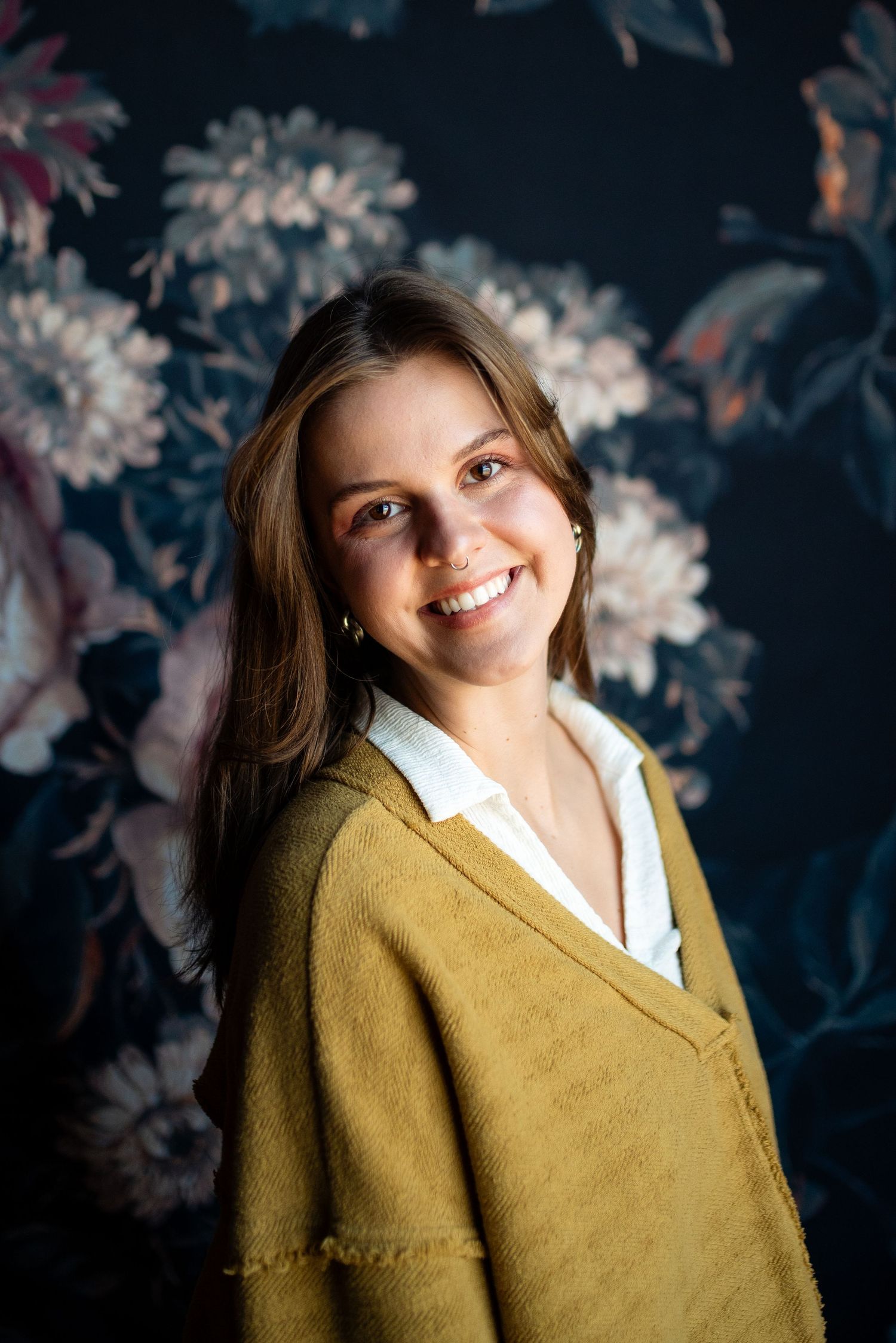 Portrait against floral wallpaper wearing mustard cardigan and white collared shirt.