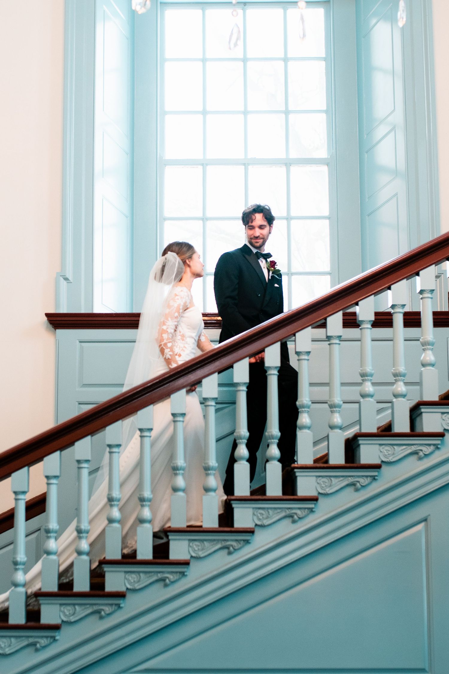 A couple in wedding attire stands on an elegant light blue staircase with white banisters in a historic building.