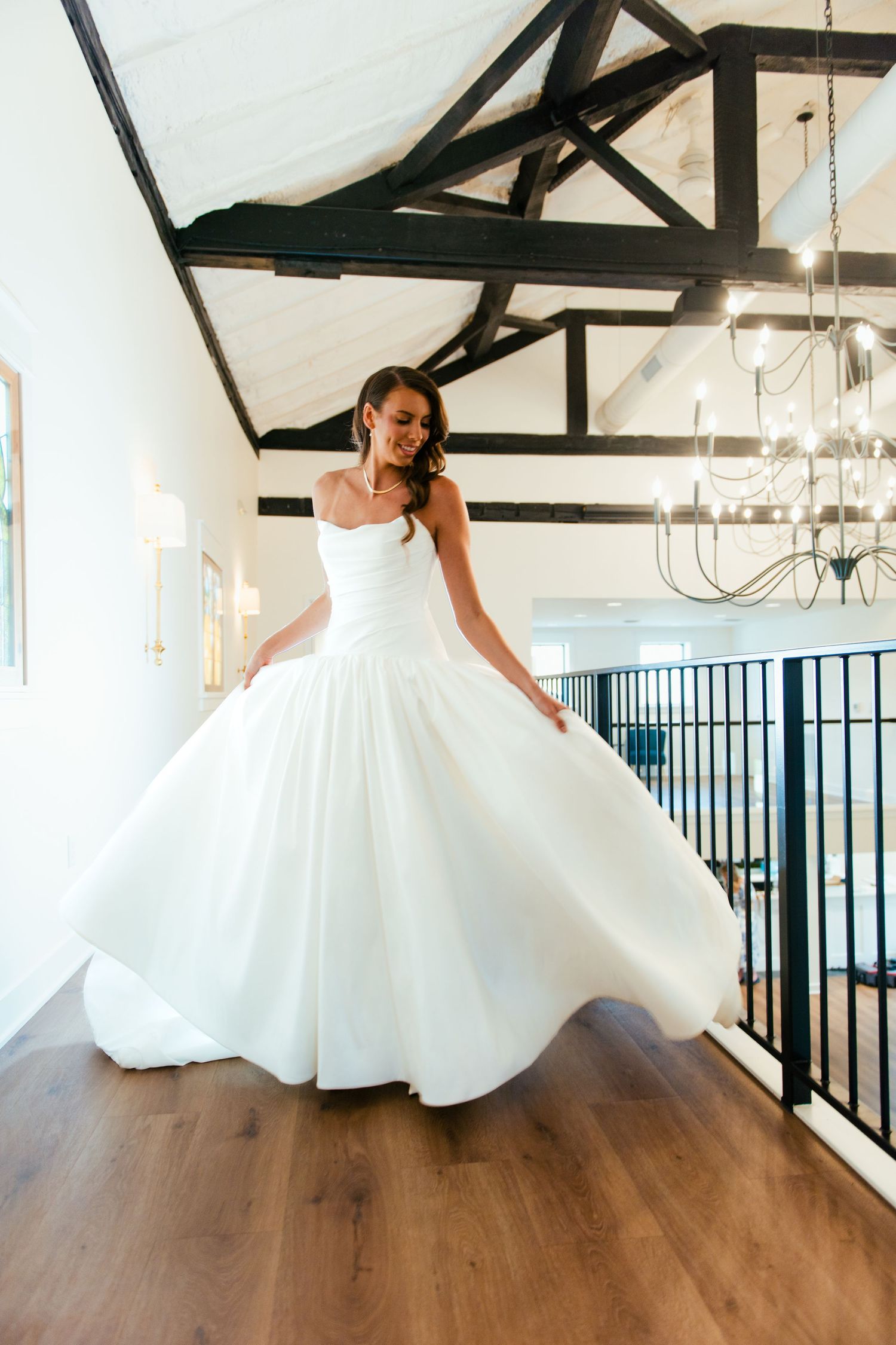 A person in a flowing white wedding dress twirls gracefully across a hardwood floor beneath exposed wooden beams.