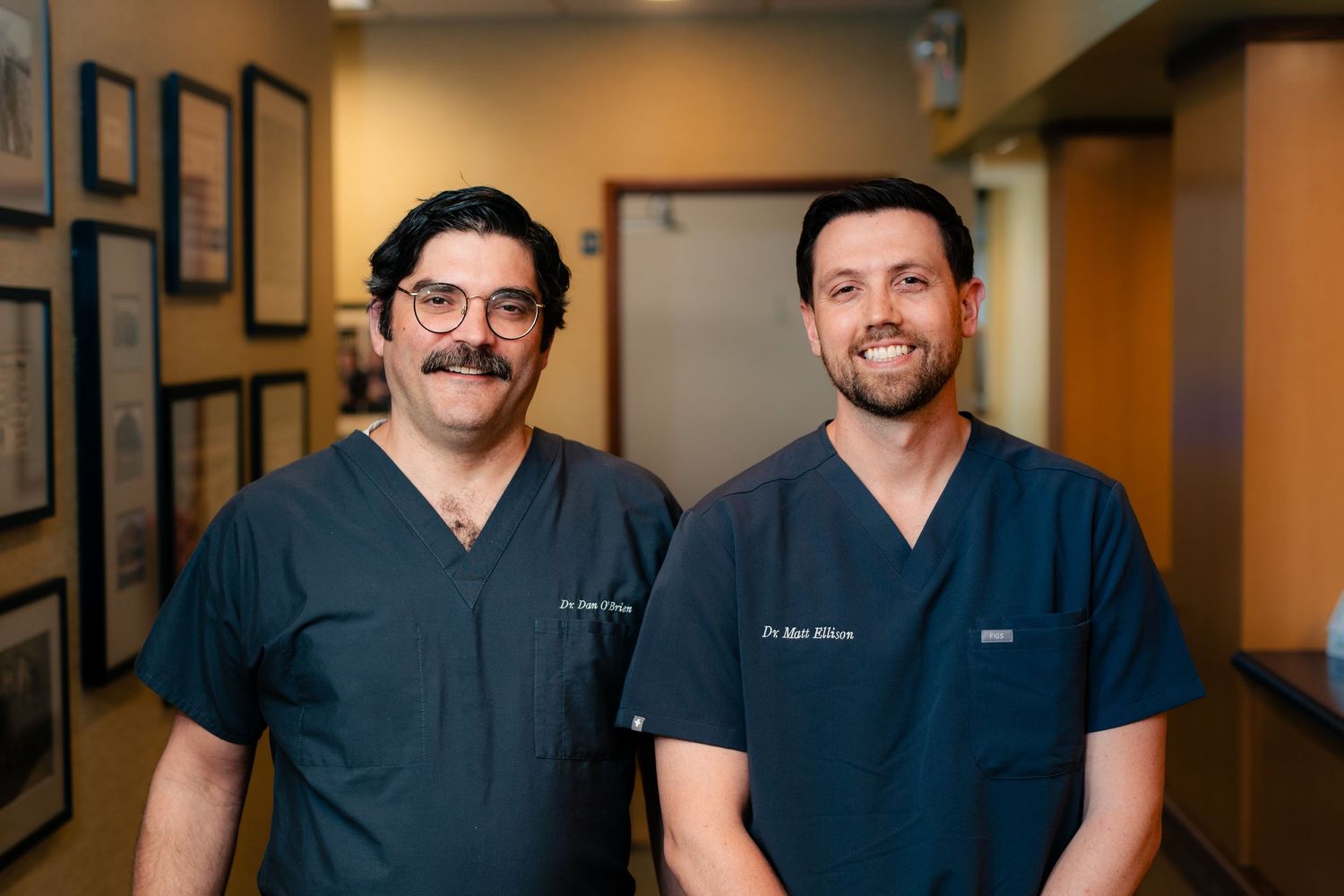 Two healthcare professionals in blue scrubs stand smiling in a medical office hallway.