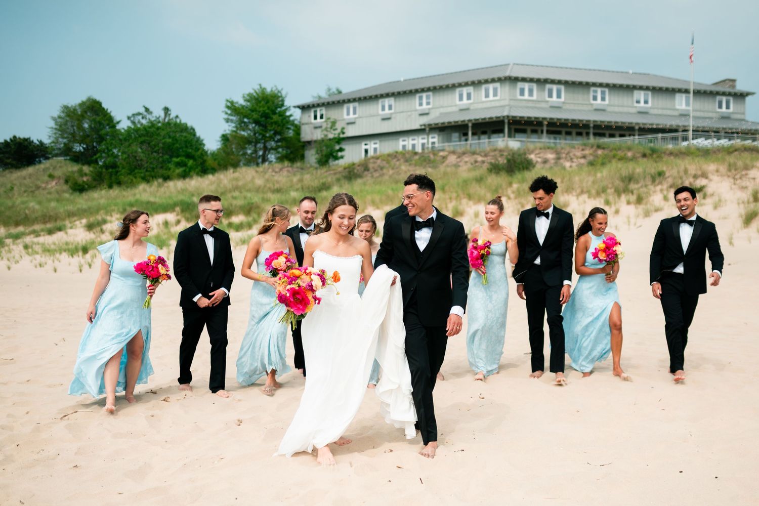 Wedding party walking on sandy beach, bridesmaids in light blue gowns, groomsmen in tuxedos, bride and groom leading.