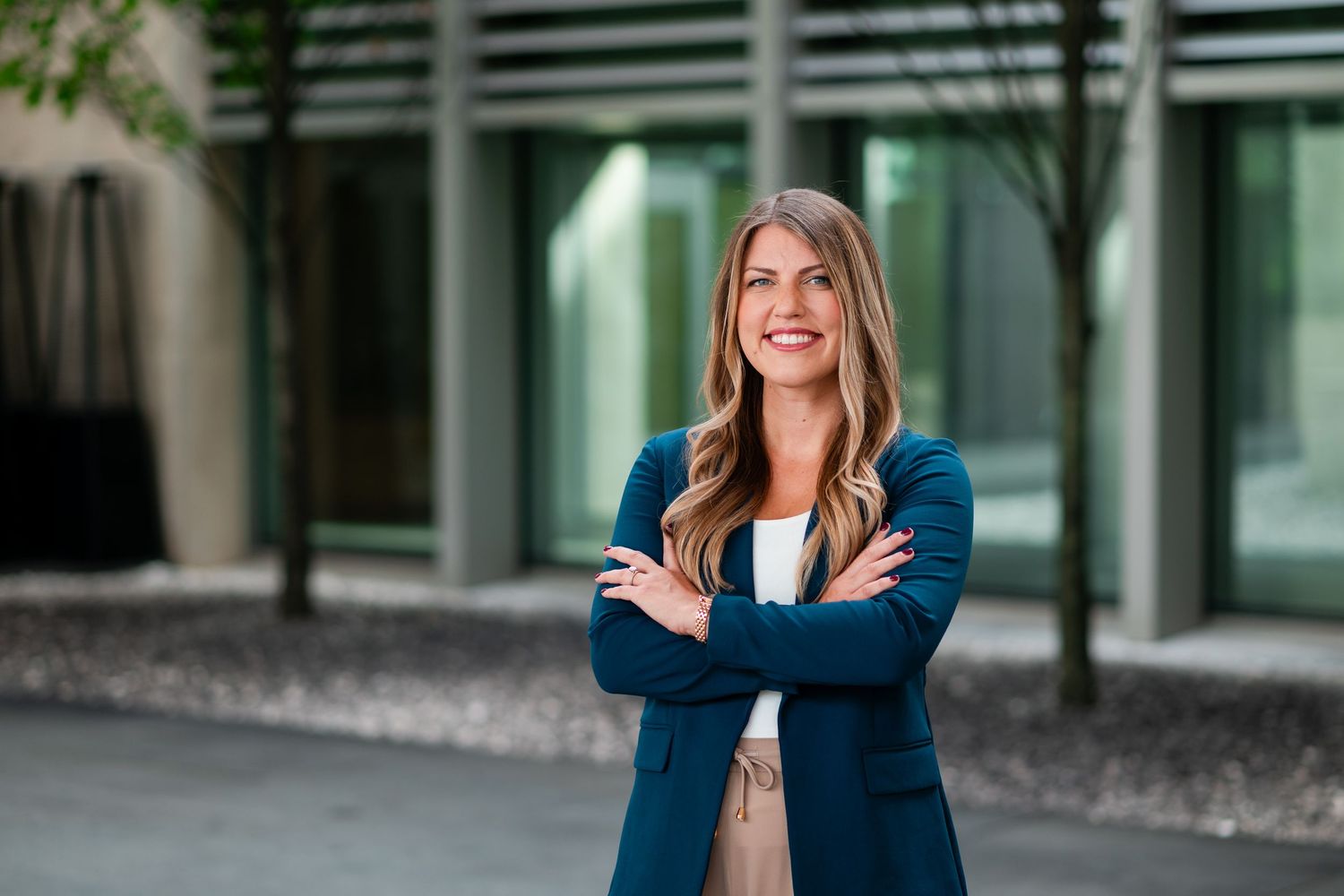 Professional woman in teal blazer smiling with arms crossed outside modern glass building.