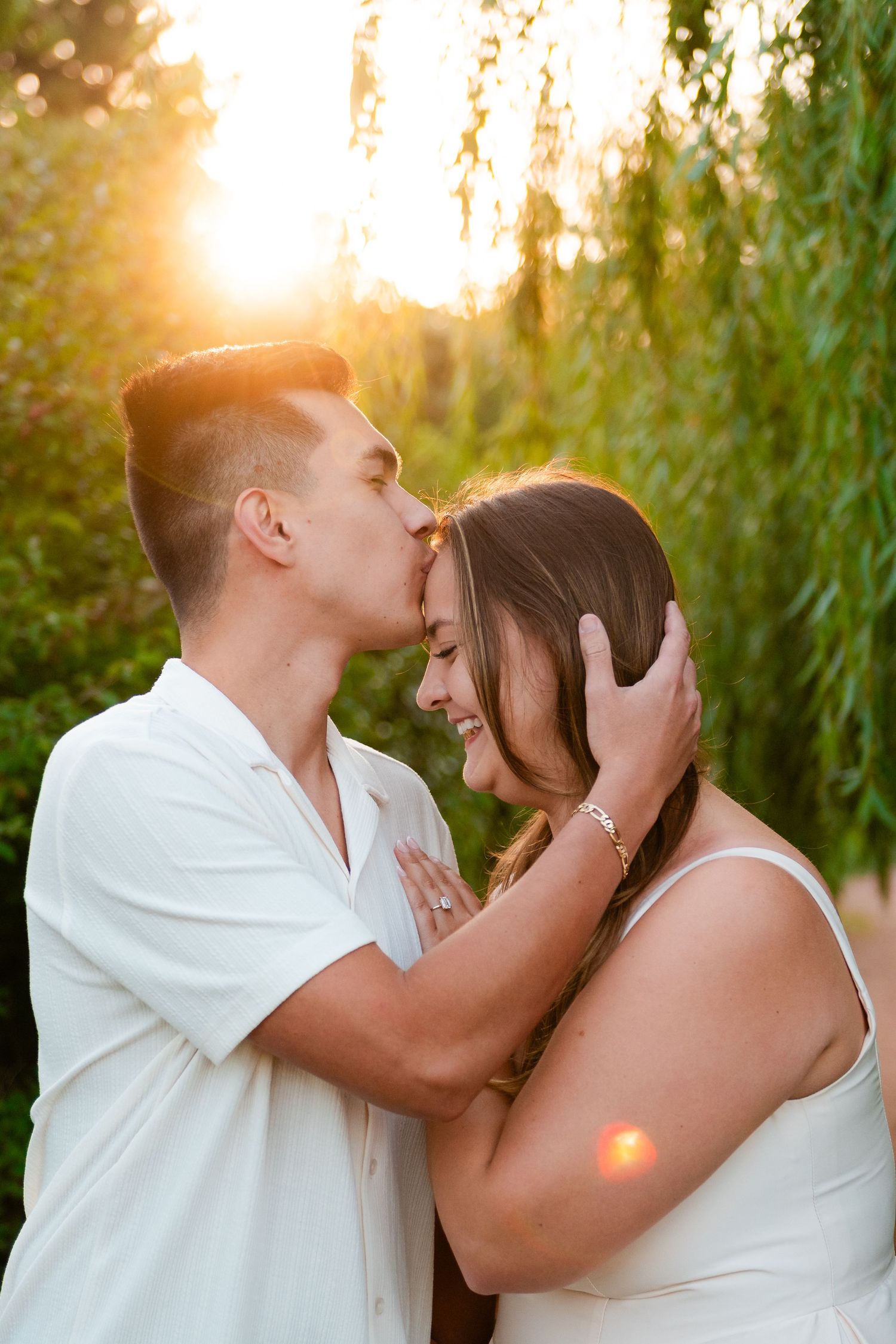 Couple sharing a tender kiss at golden hour sunset, surrounded by lush green weeping willow trees.