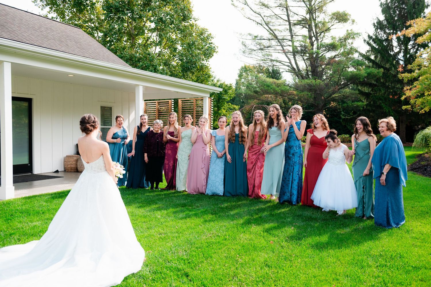 Bride in white gown reveals wedding dress to bridesmaids in colorful dresses on green lawn outside white venue.