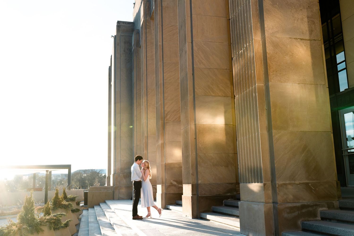Couple sharing a romantic moment beside grand golden columns of a stunning architectural building at sunset.
