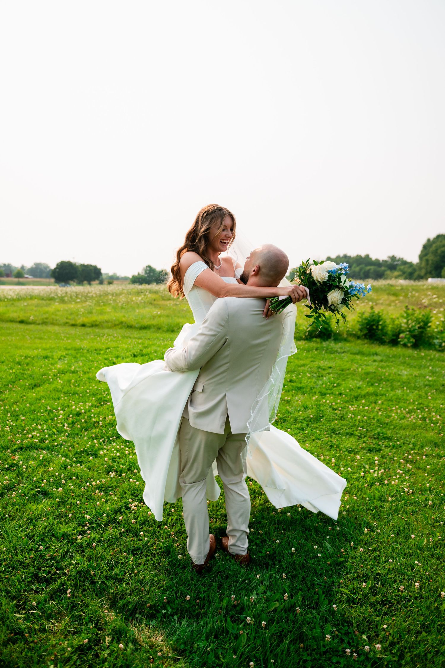 Groom lifting bride in white dress on green field, both smiling, bride holding blue and white bouquet.
