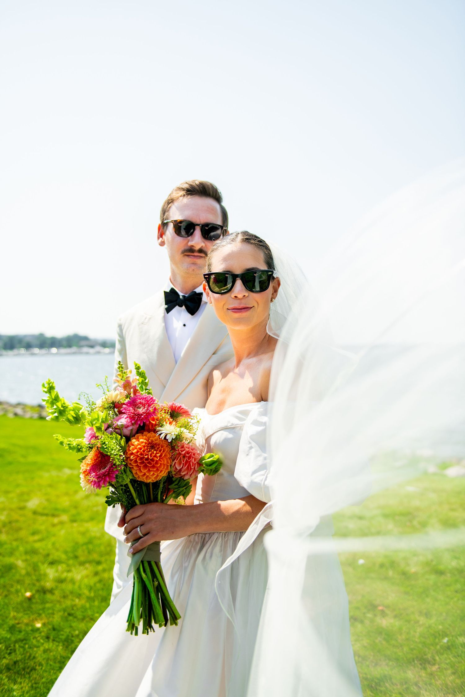 Bride and groom in white attire and sunglasses pose outdoors by waterfront, bride holding vibrant orange and pink bouquet.