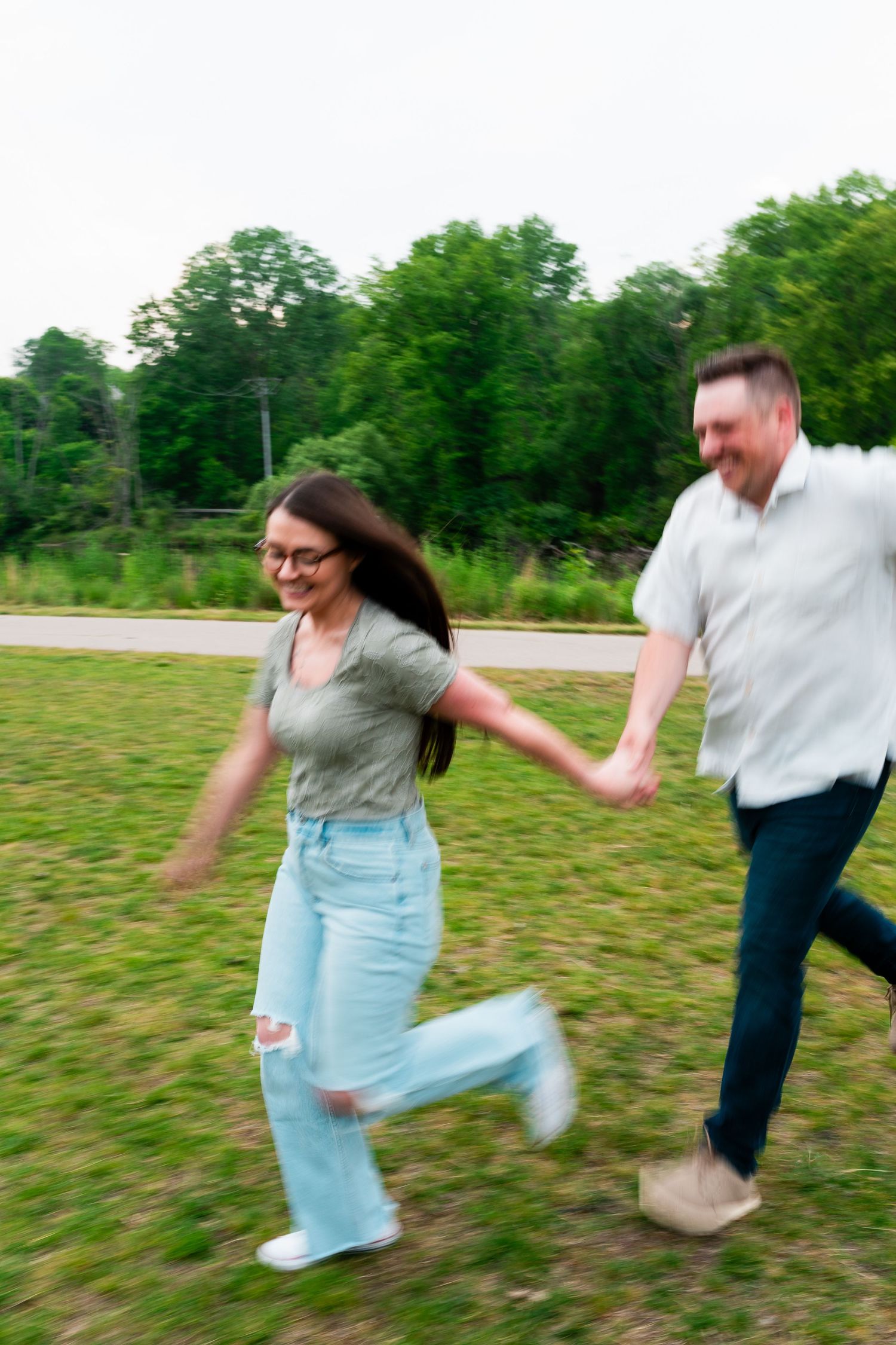 Couple running and holding hands in a grassy field with trees in the background on a sunny day.