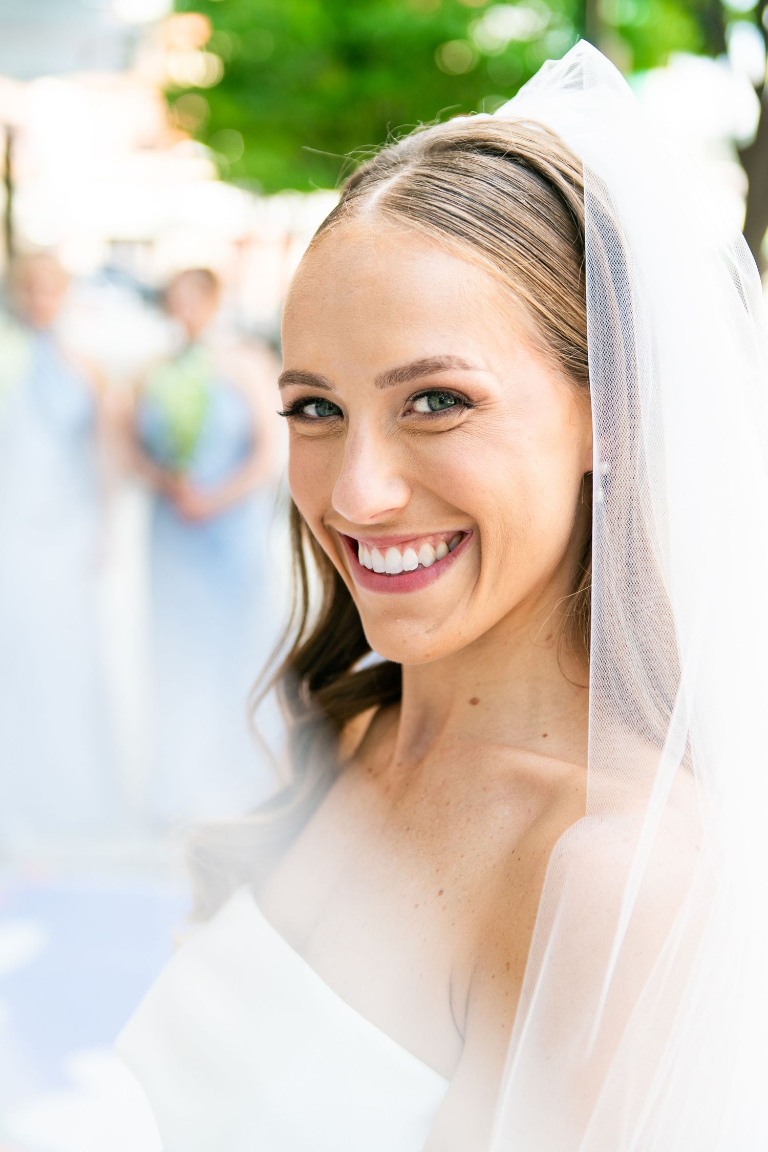 A radiant bride wearing a white veil smiles joyfully on her wedding day with soft natural lighting and greenery in background.
