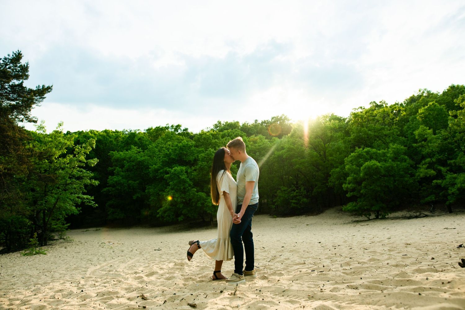 Couple embraces on sandy beach surrounded by green forest while sun creates magical glow in background.