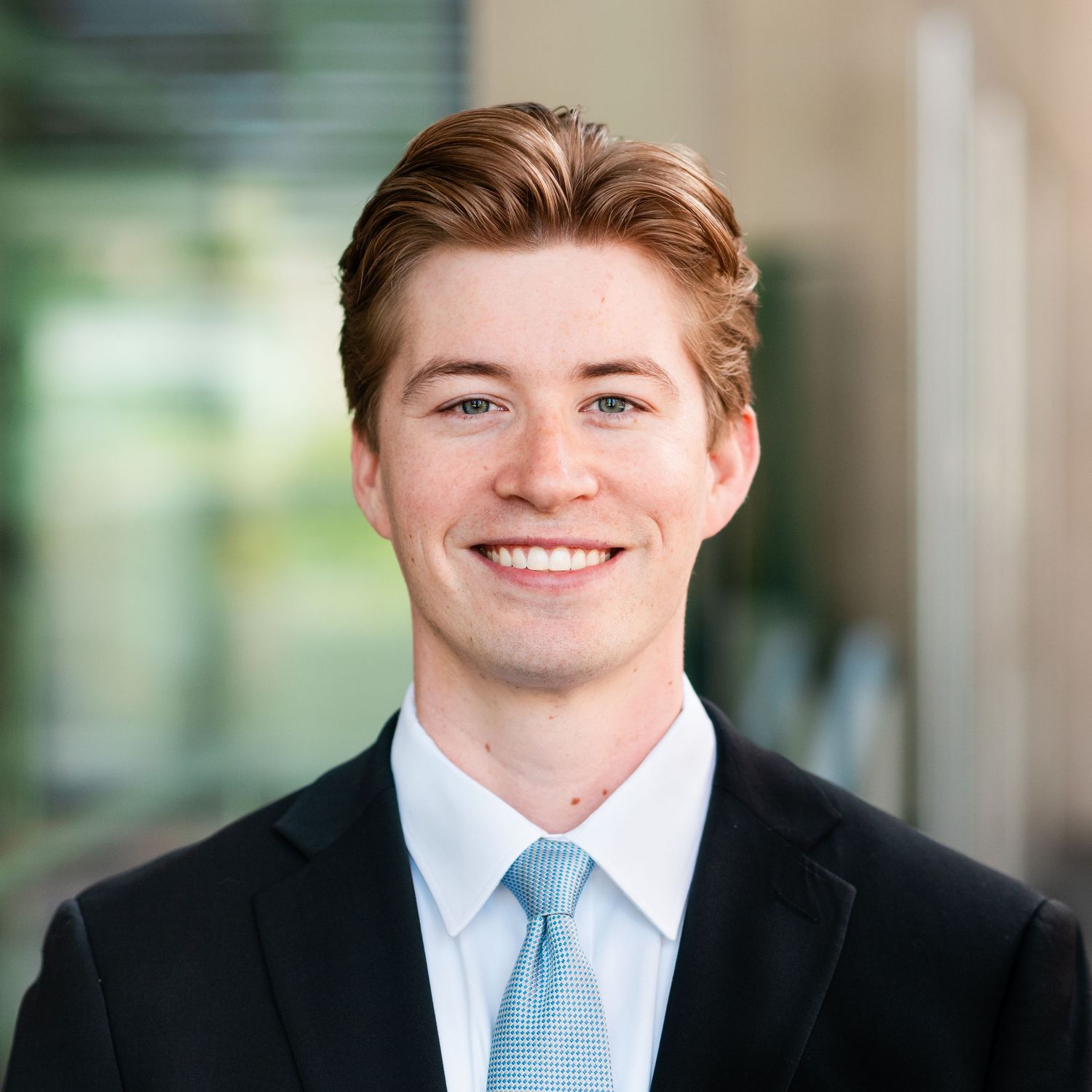 Professional headshot portrait of someone wearing a light blue dress shirt and tie against a blurred office background.