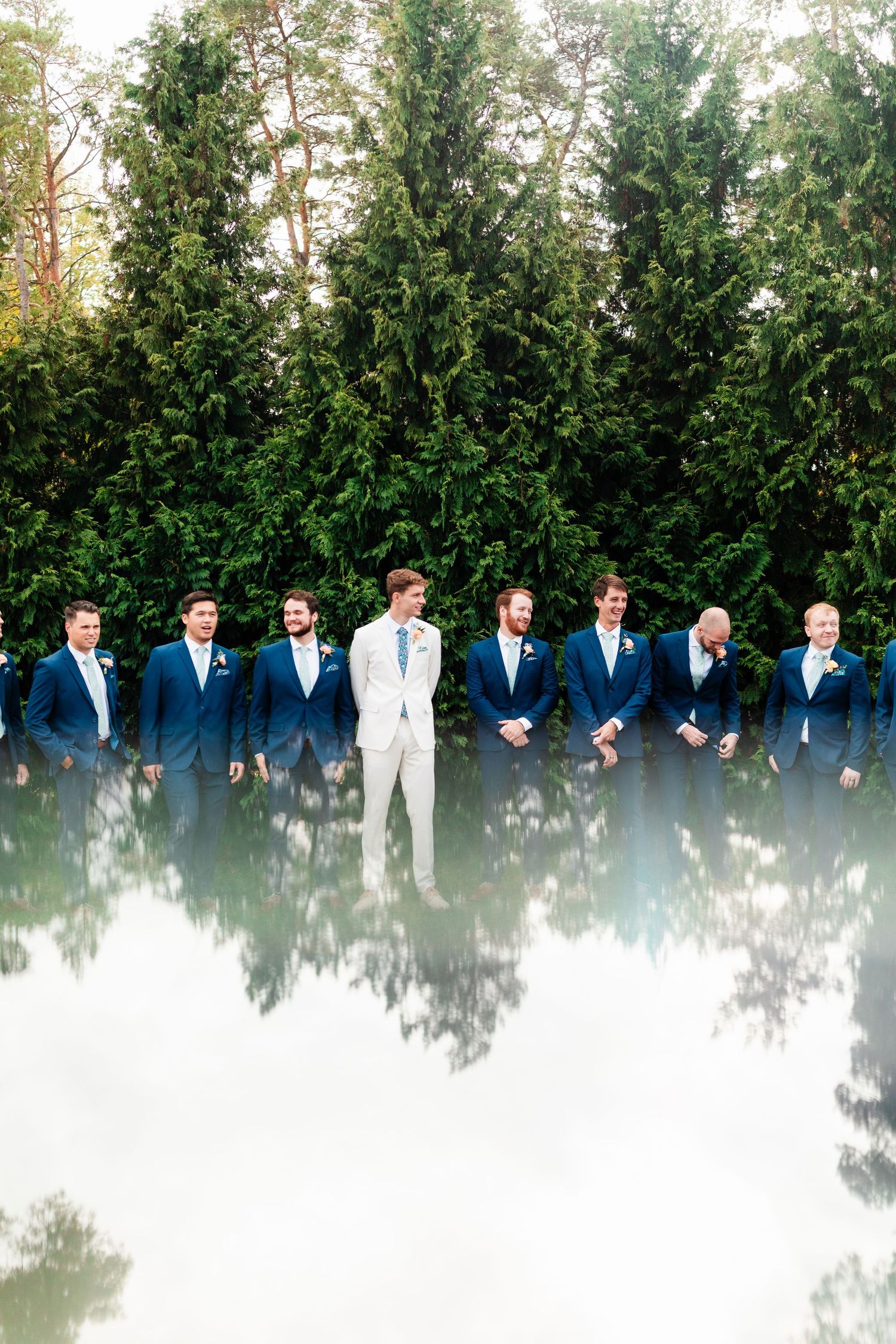 A group of groomsmen in navy suits and one in white stand in a line against a backdrop of green trees in a grand rapids wedding