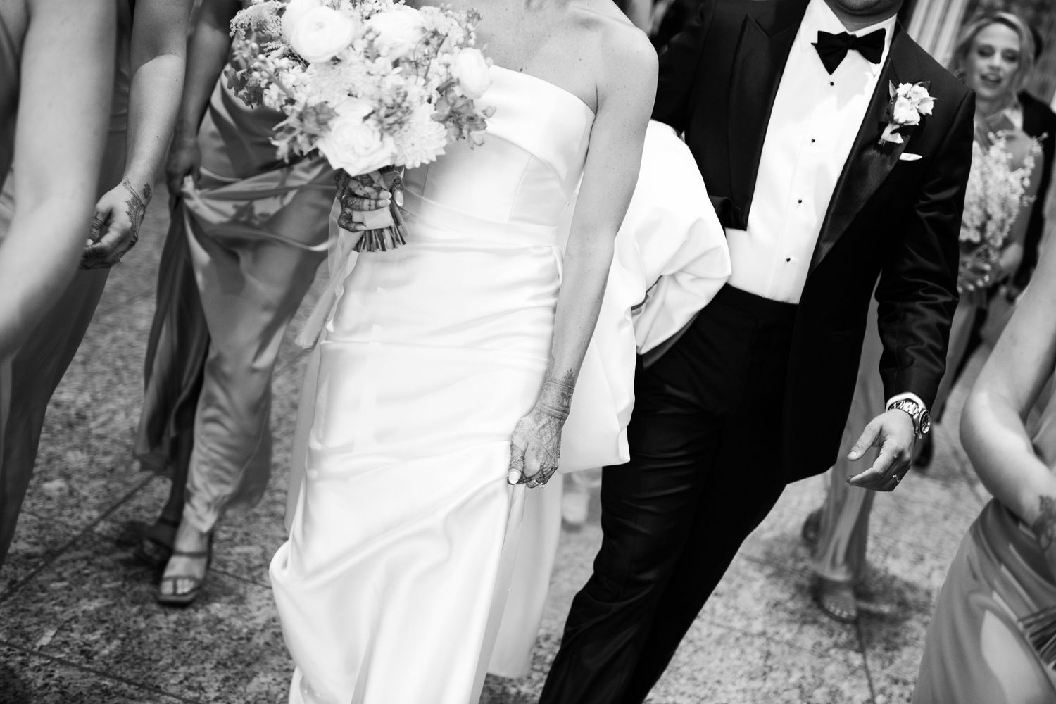 Black and white photo of bride in strapless gown holding bouquet walking with groom in tuxedo surrounded by wedding party.