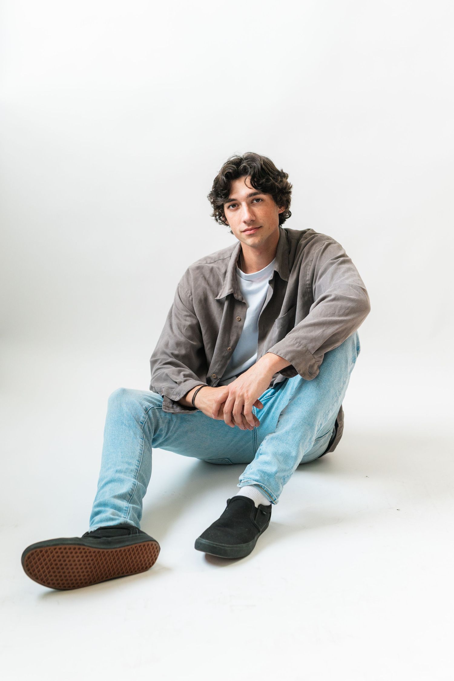 Young man with curly hair sitting on white background wearing gray jacket, blue jeans, and black sneakers.