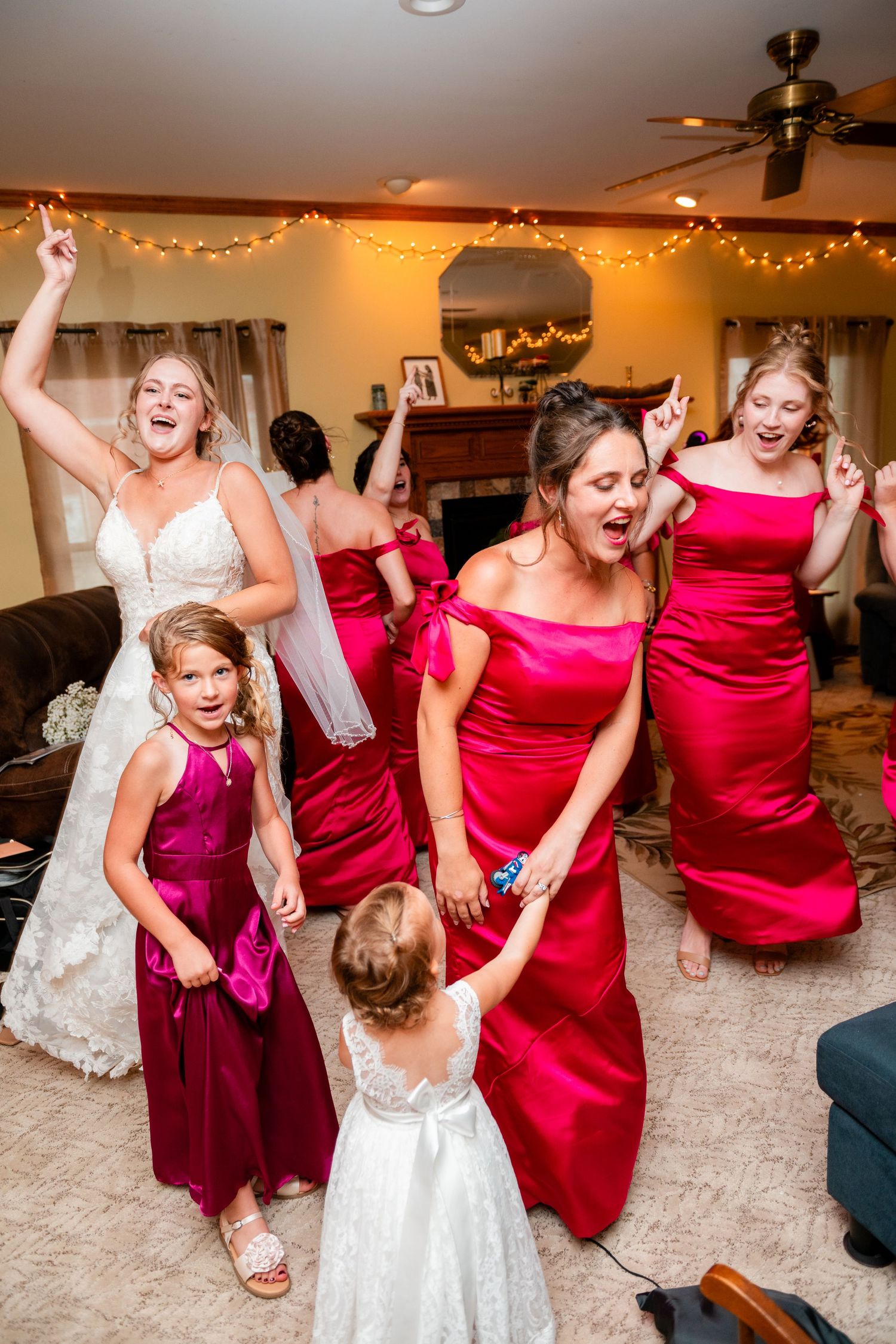 A group in red bridesmaid dresses and a white wedding gown dance energetically at a reception party.