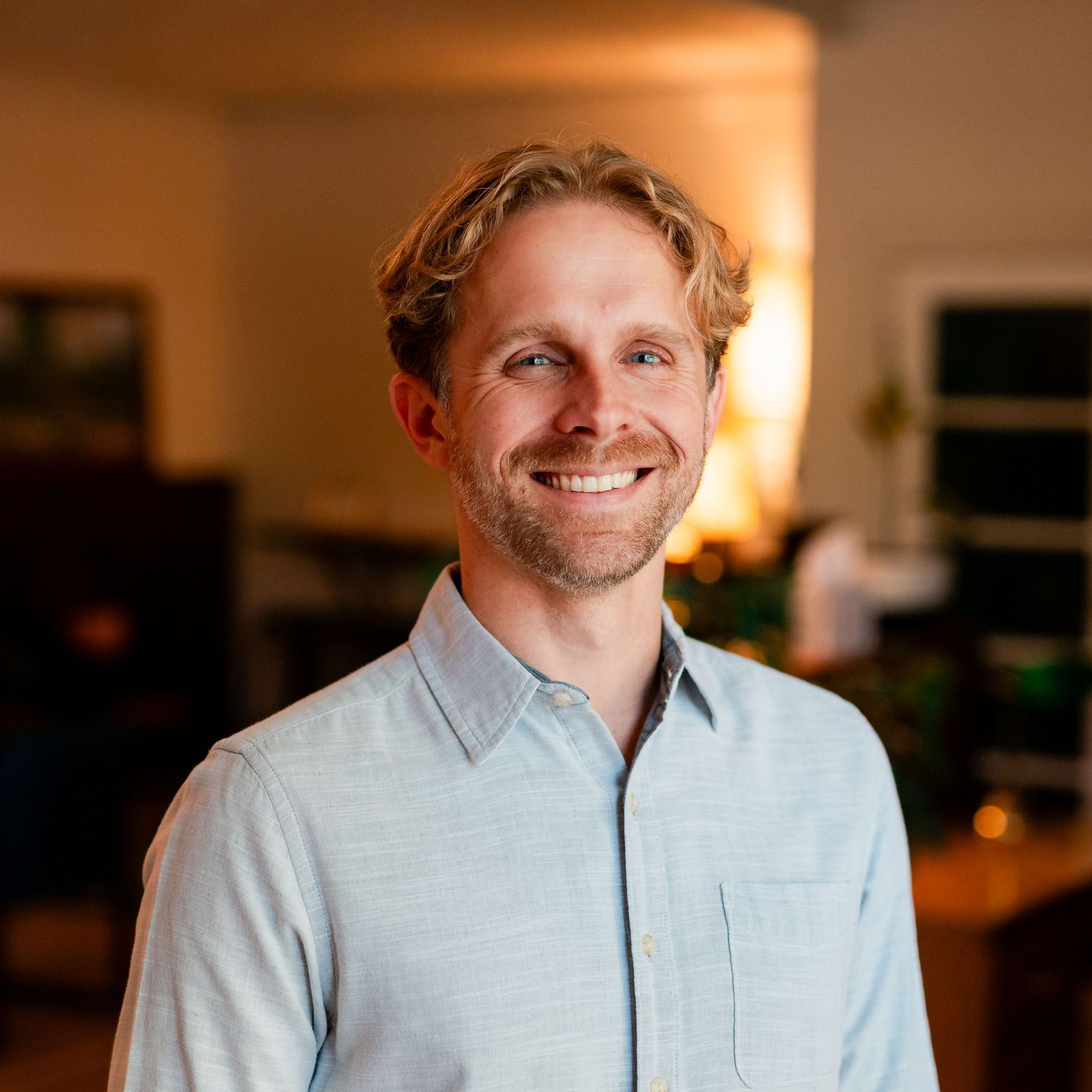 Professional headshot of someone in a light blue button-down shirt against a warm-toned blurry background.