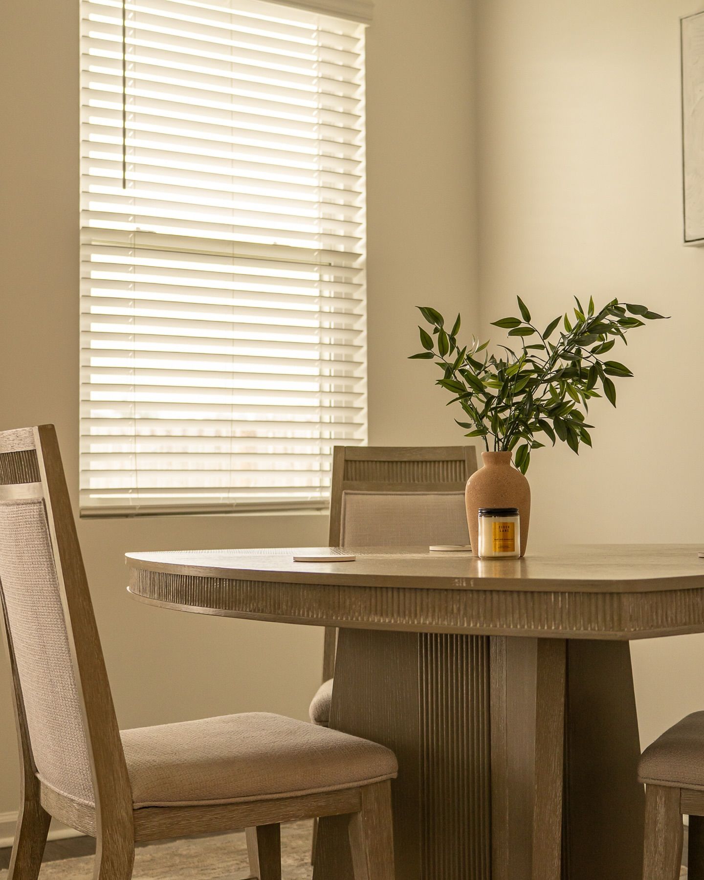 Minimalist dining space with round wooden table and beige chairs next to window with blinds for real estate photography in Augusta GA.