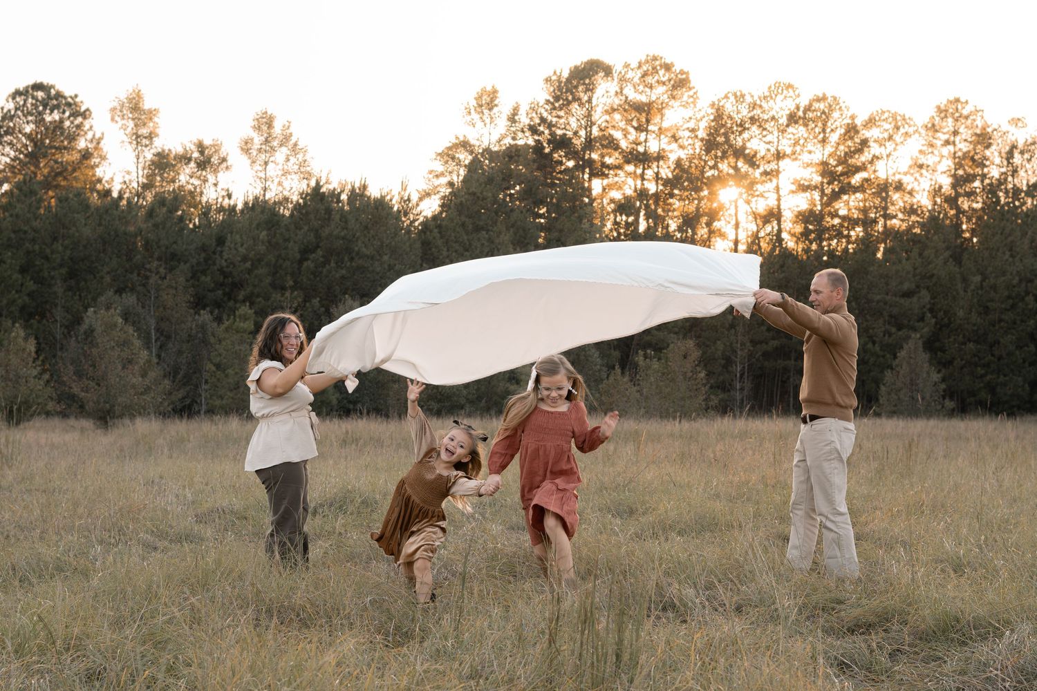 Family playing with white sheet in golden field at sunset while children run underneath in Evans, GA photoshoot.