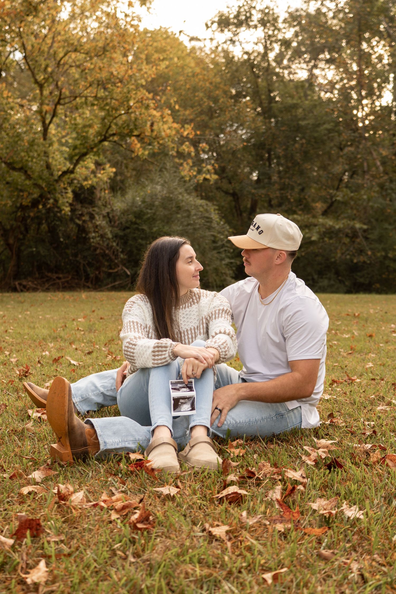 A couple relaxes together on autumn ground covered in fallen brown leaves during a romantic outdoor photoshoot.