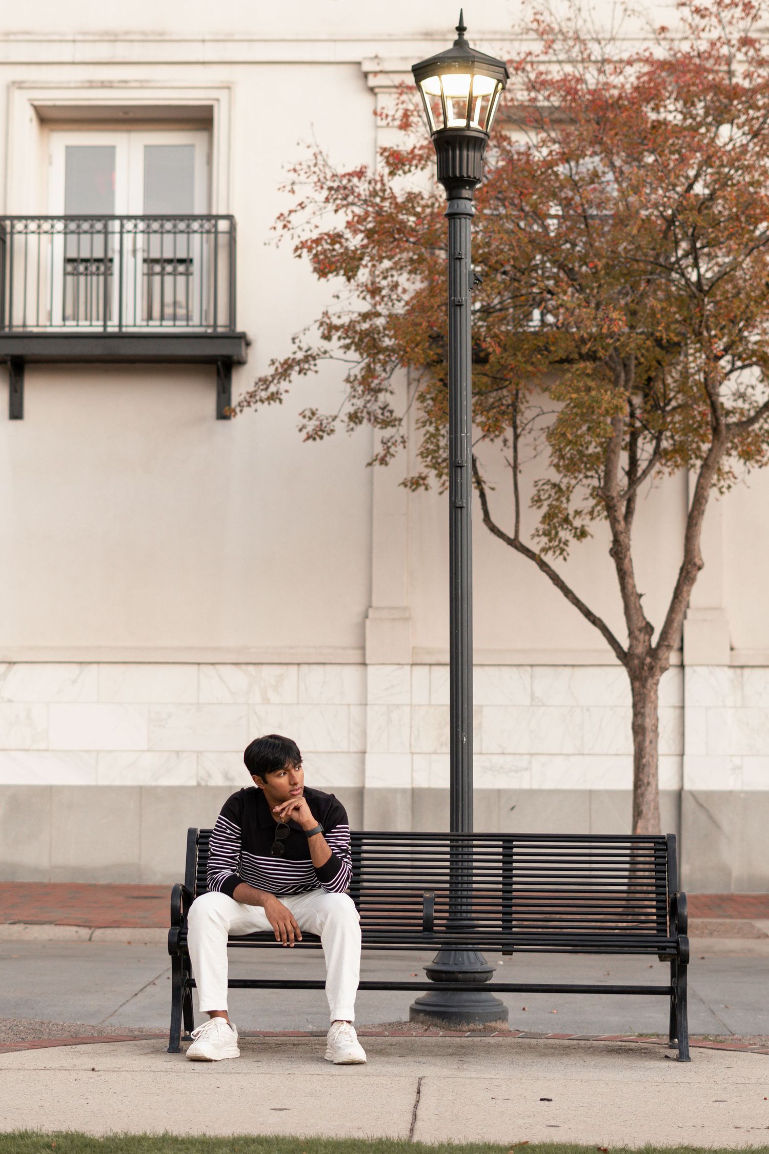 A person in dark clothing sits alone on a public bench near a white building and street lamp in autumn.