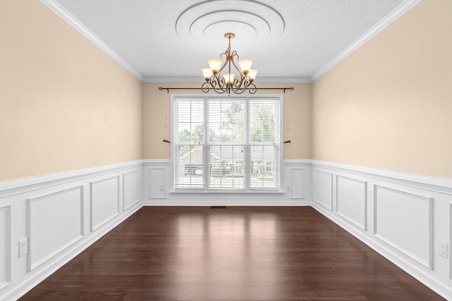 Elegant dining room with white wainscoting, beige walls, and a crystal chandelier hanging from the tray ceiling for real estate photography.
