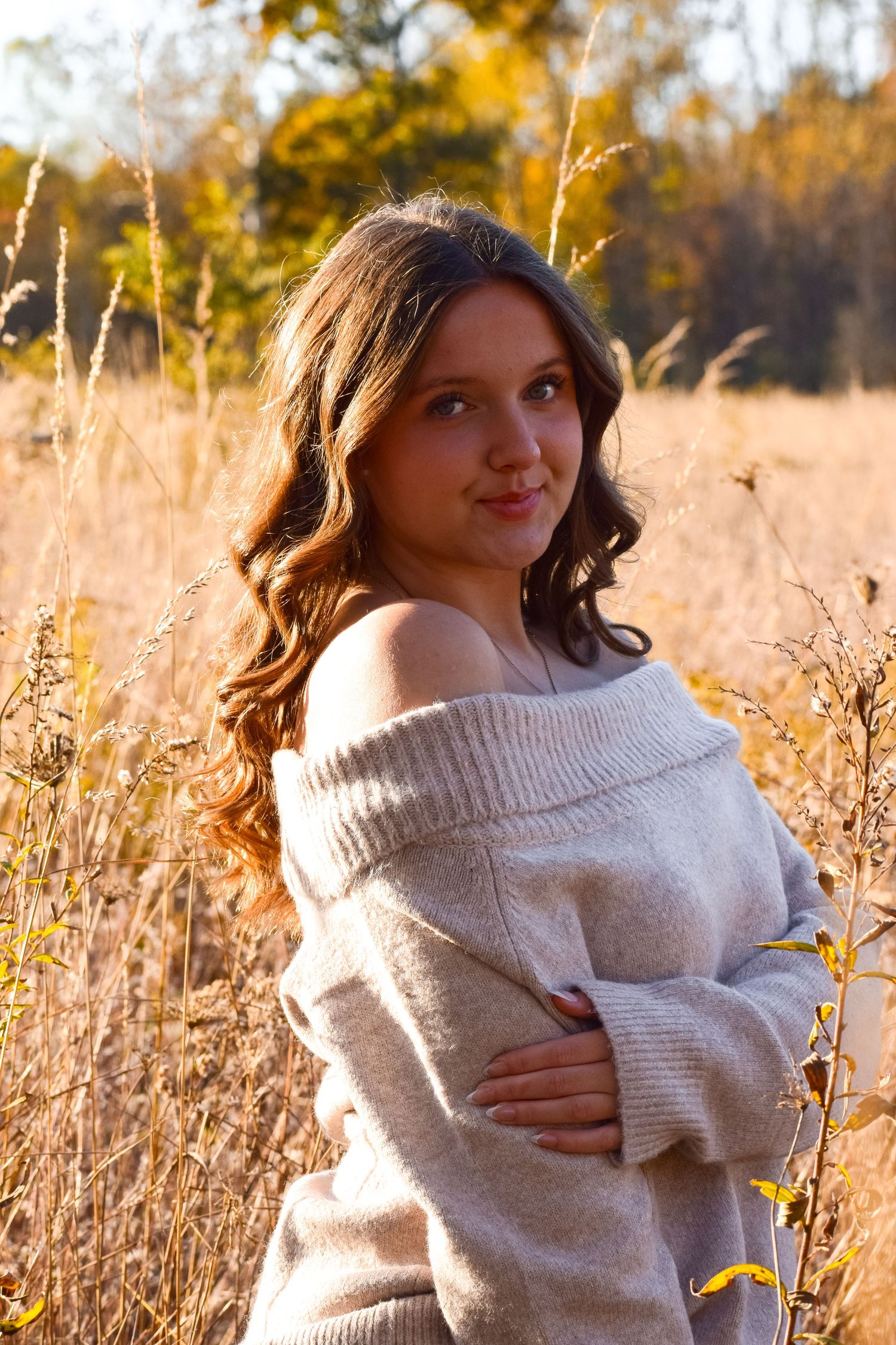 Woman in beige sweater posing in golden autumn field during sunset.