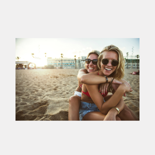 Friends enjoying a sunny day at the beach, laughing and embracing while wearing sunglasses.