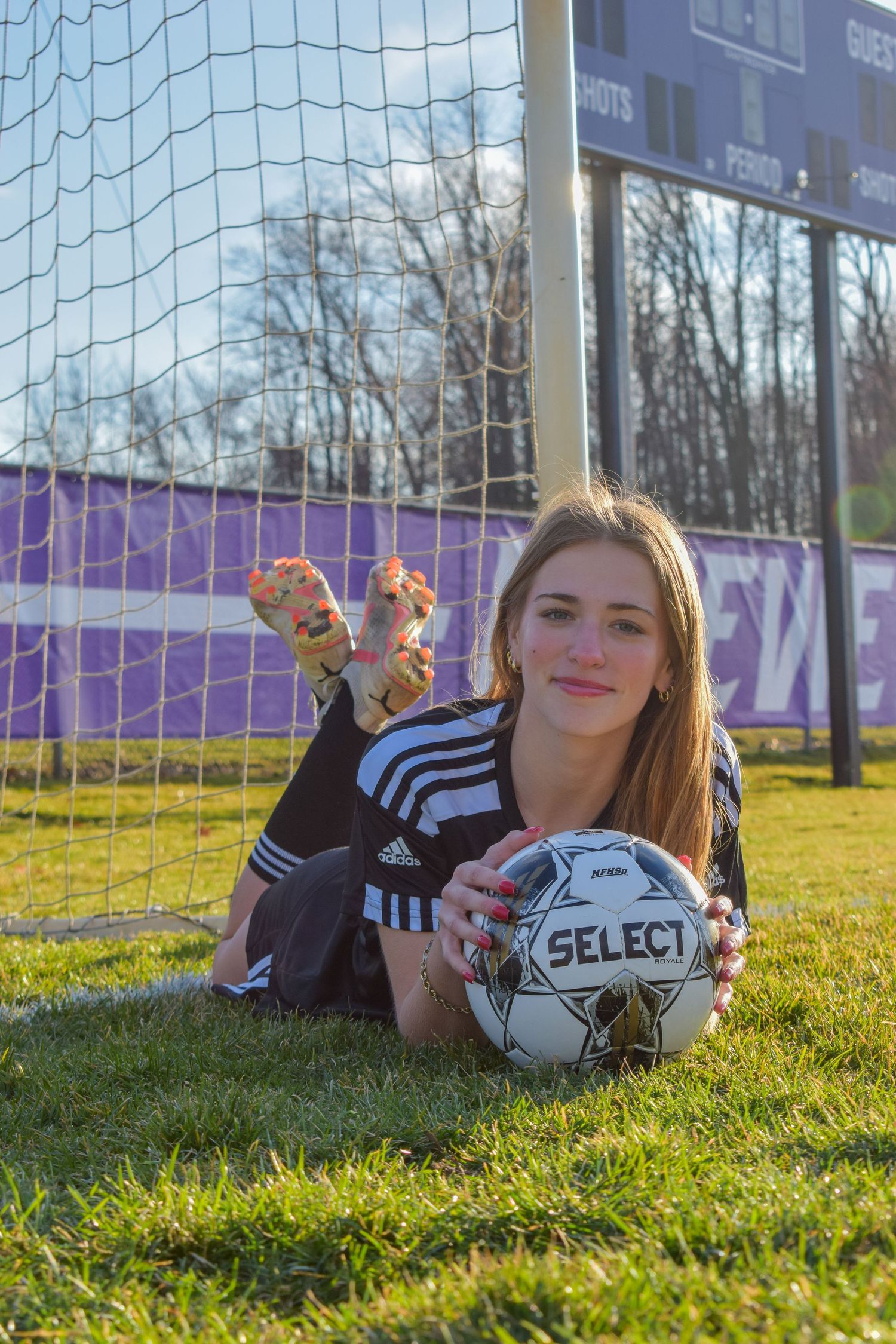 Soccer player lying on grass by goal post with Select ball and purple backdrop.