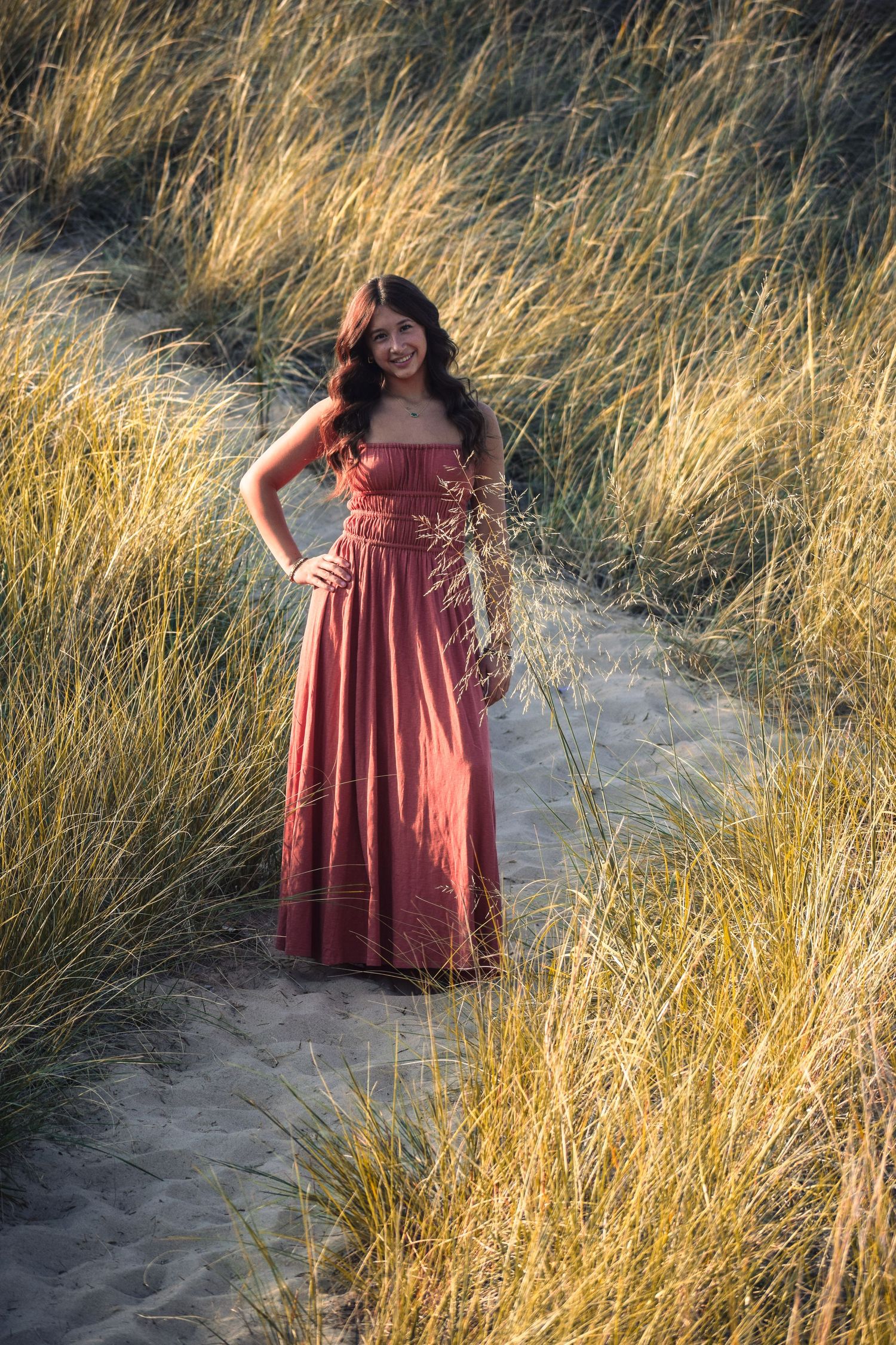 Figure in rust-colored dress poses along path through golden grass at sunset.