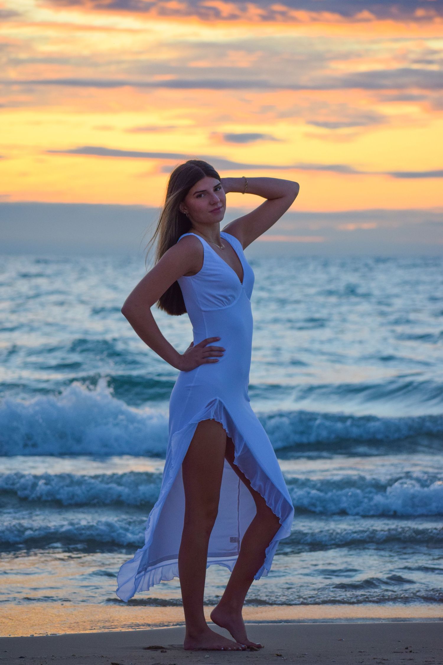 Figure in white dress poses dynamically against dramatic orange sunset sky and crashing waves.