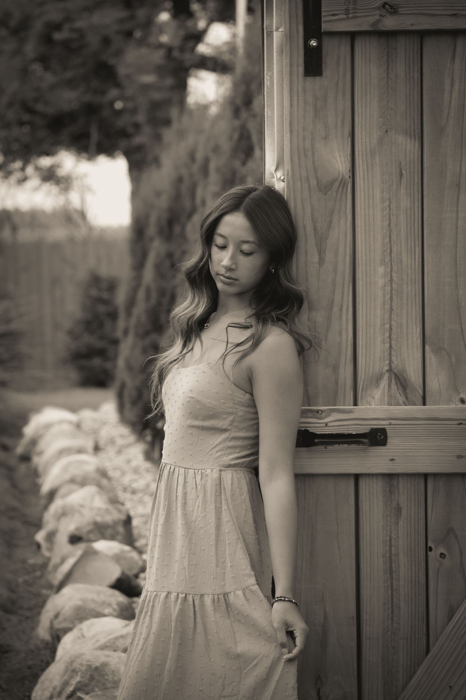 Woman in light dress poses thoughtfully by wooden door in black and white photograph.