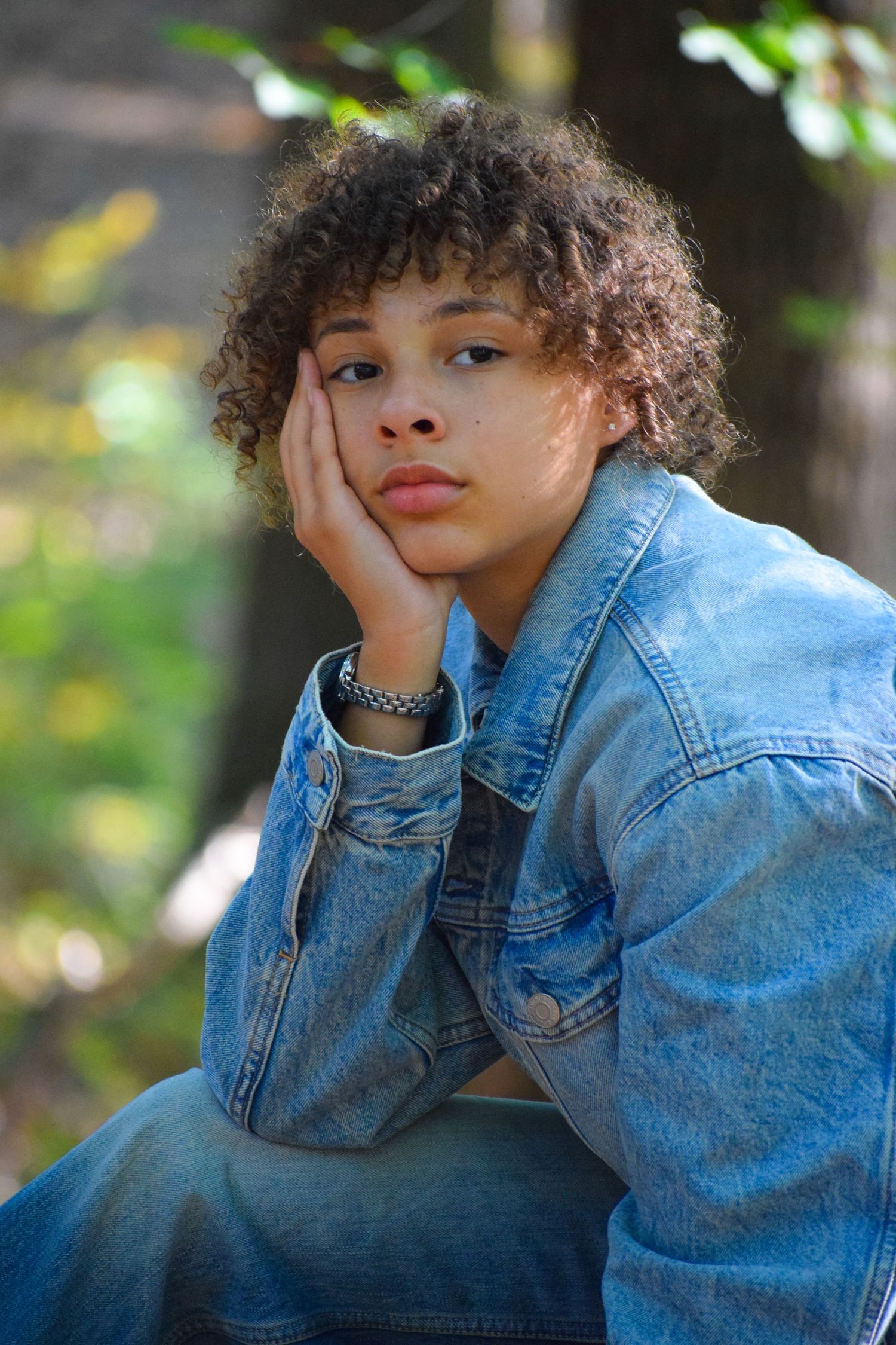 Person in denim jacket poses thoughtfully against blurred natural background.