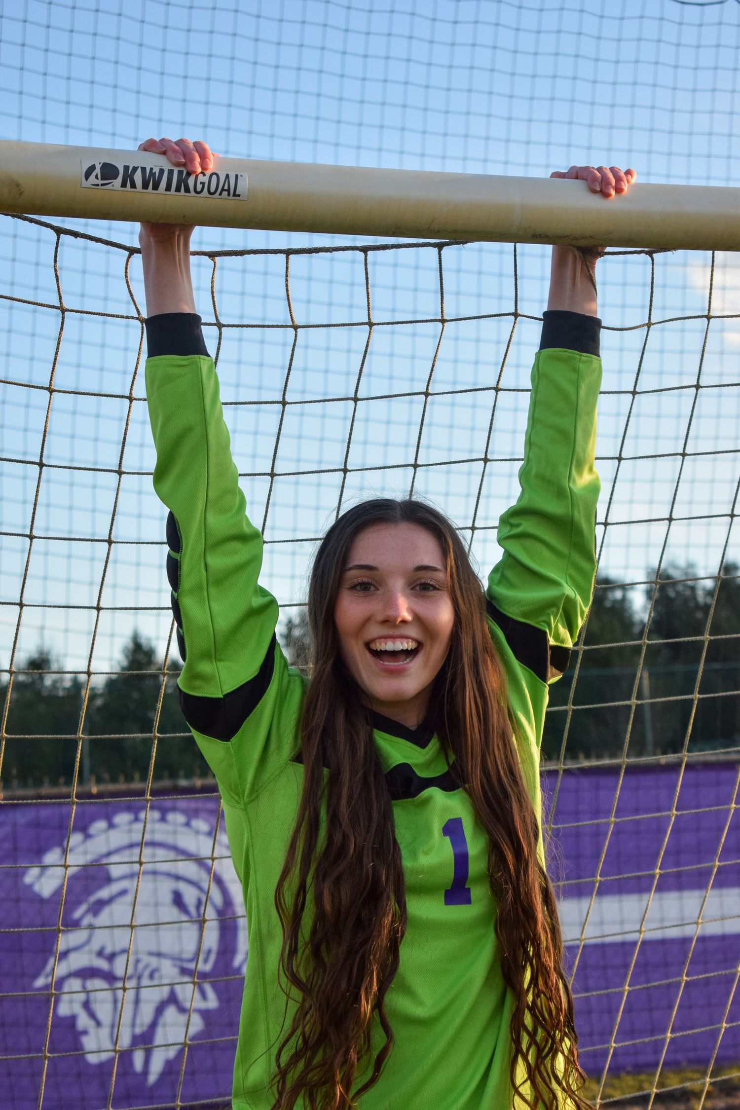 Soccer goalkeeper in bright green jersey hanging from goal crossbar.