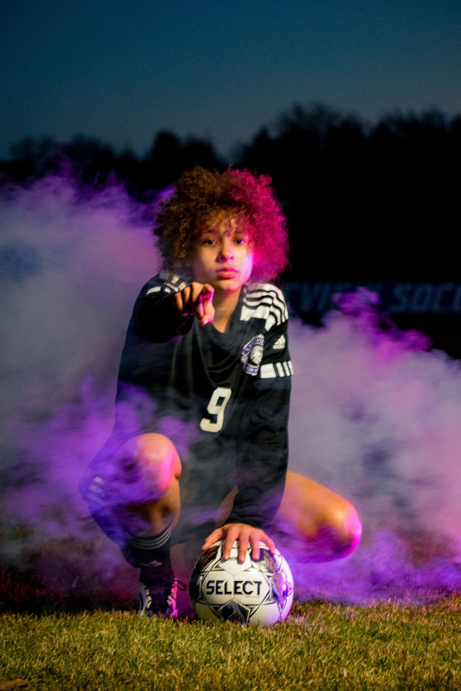 Soccer player lying on grass with Select brand ball surrounded by colorful smoke.