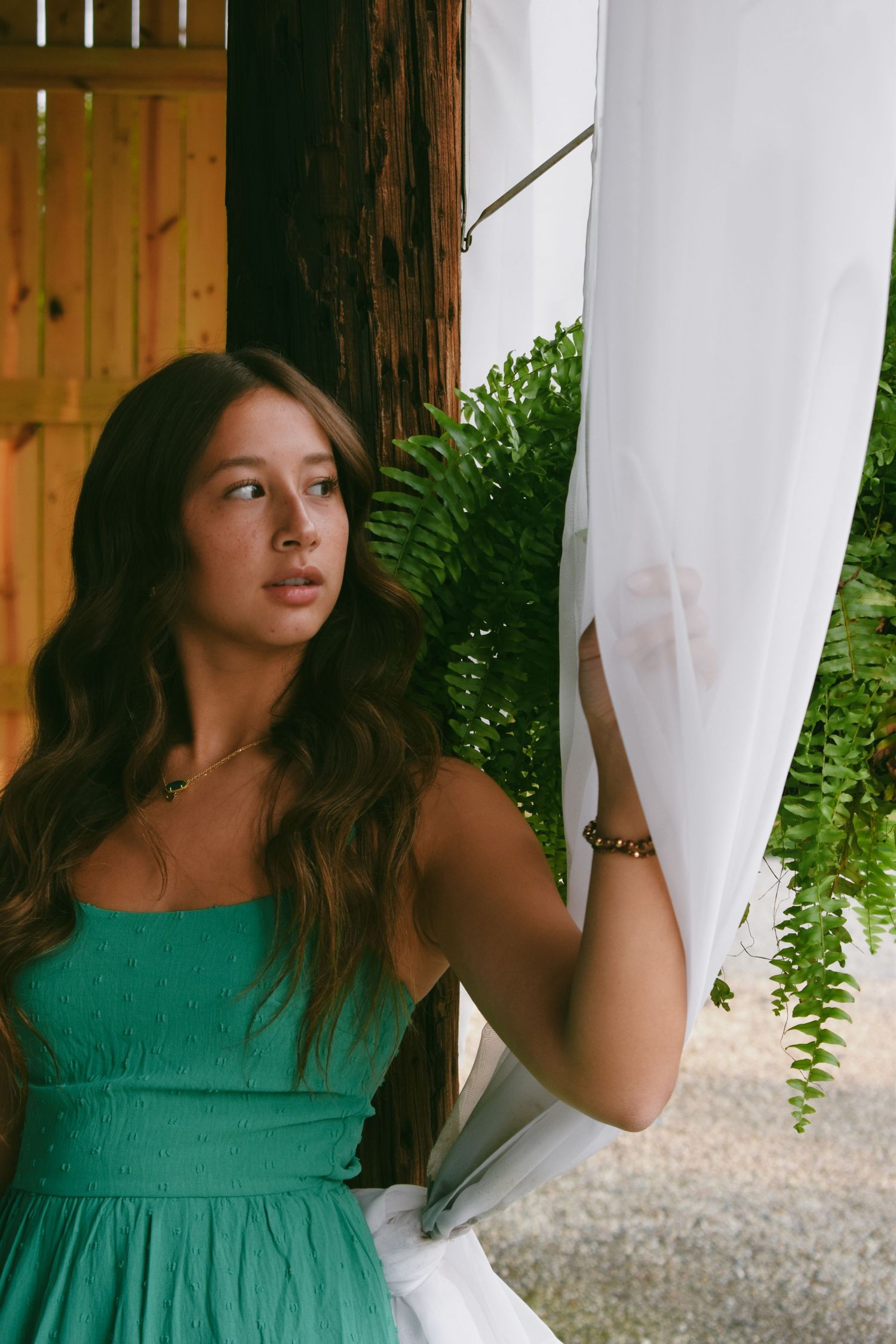 Woman in emerald green dress poses near white curtains and ferns in natural lighting.