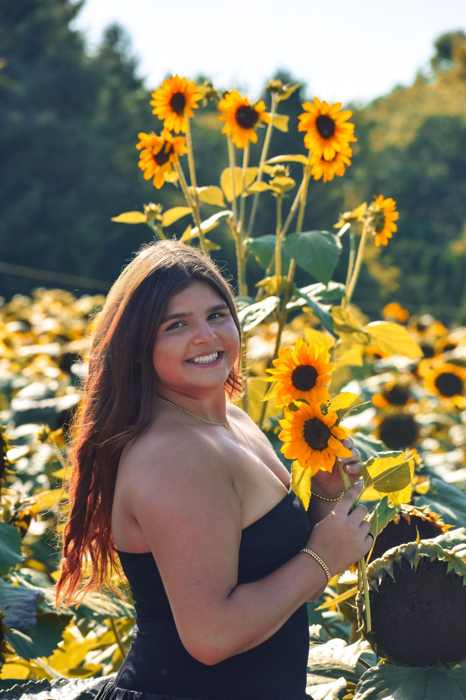 Woman in black dress smiles while holding sunflowers in blooming garden.