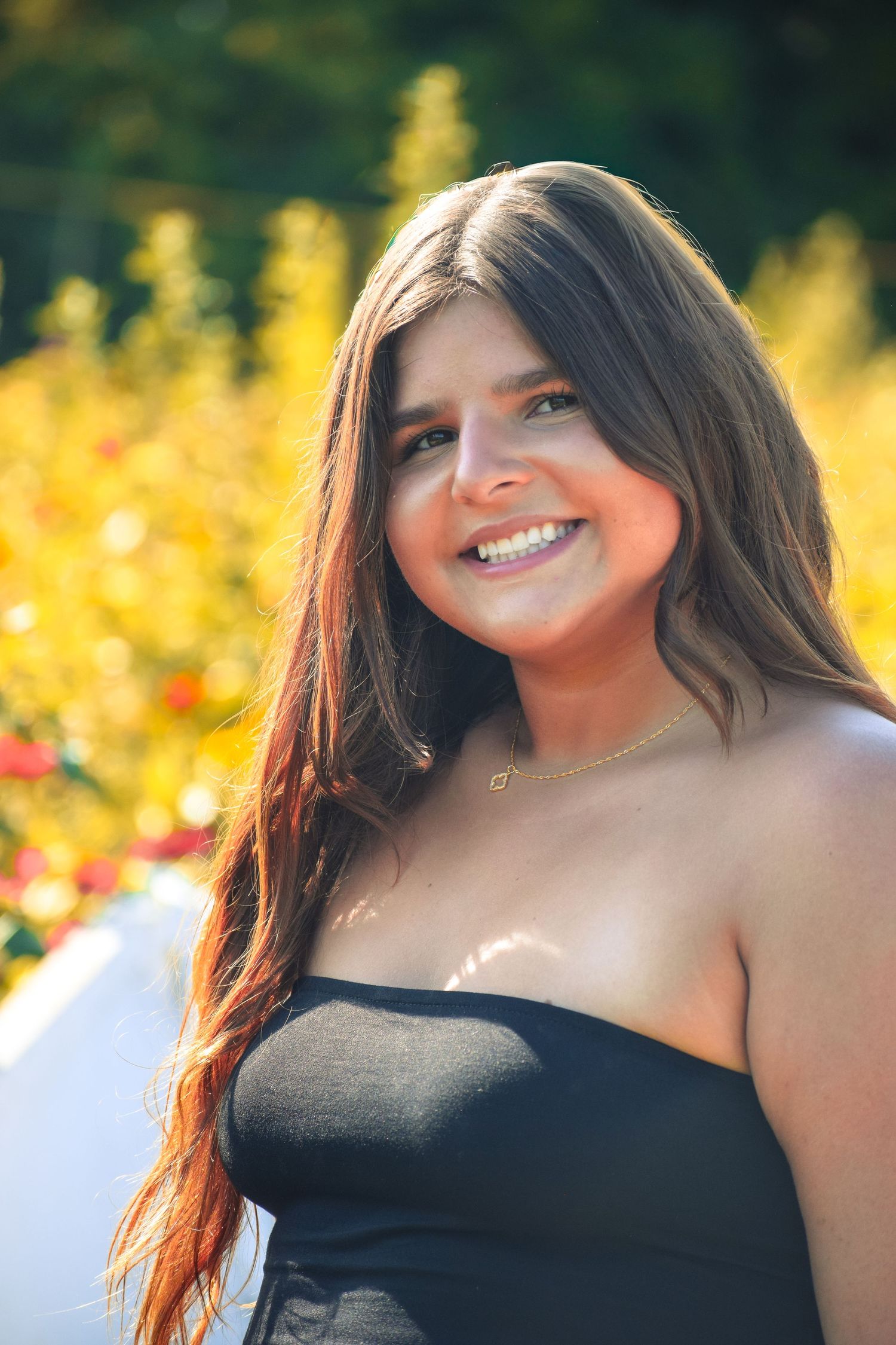 Woman in black top smiles brightly against yellow flowers in summer garden.