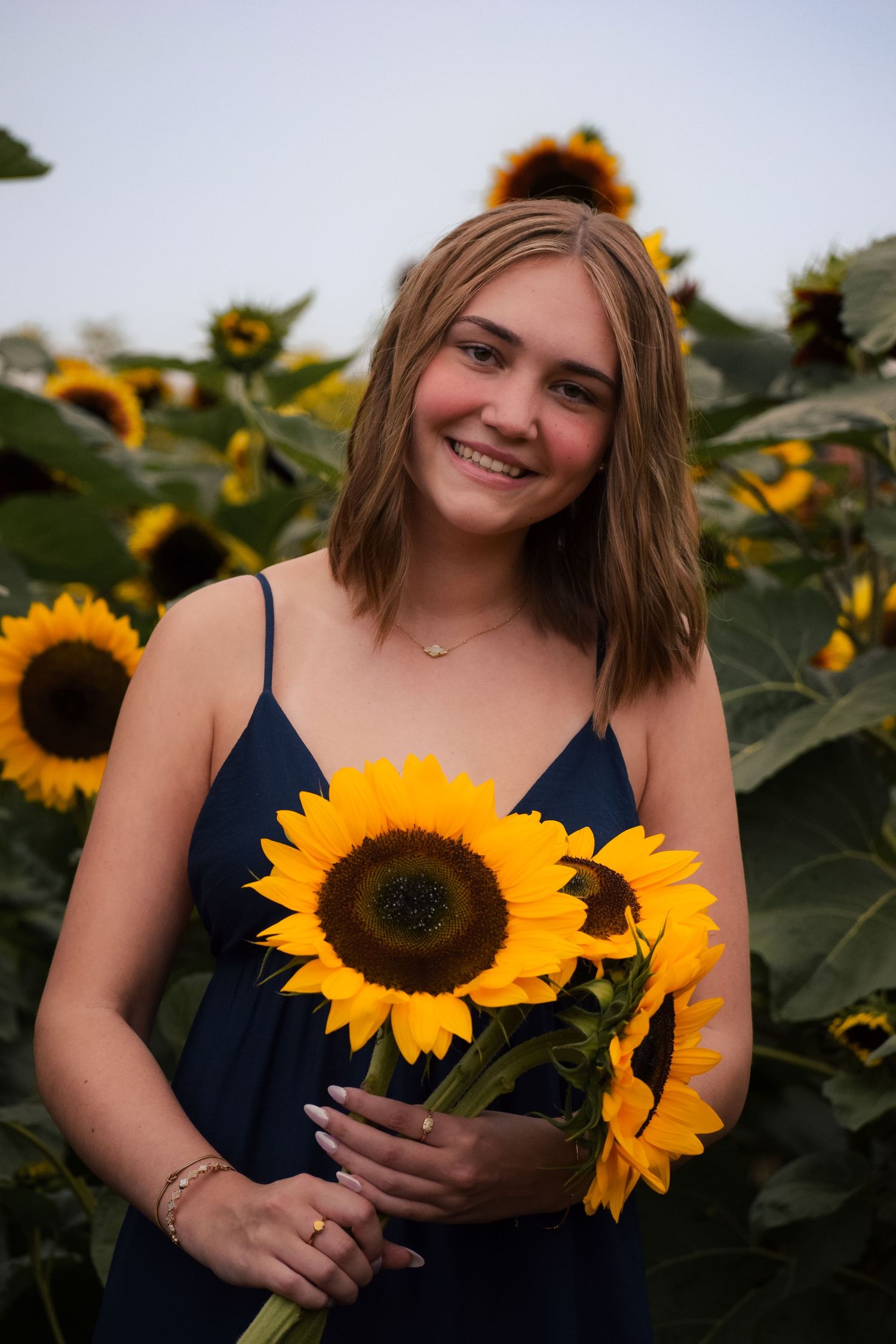Smiling young woman in navy dress holding sunflowers in a vibrant sunflower field.