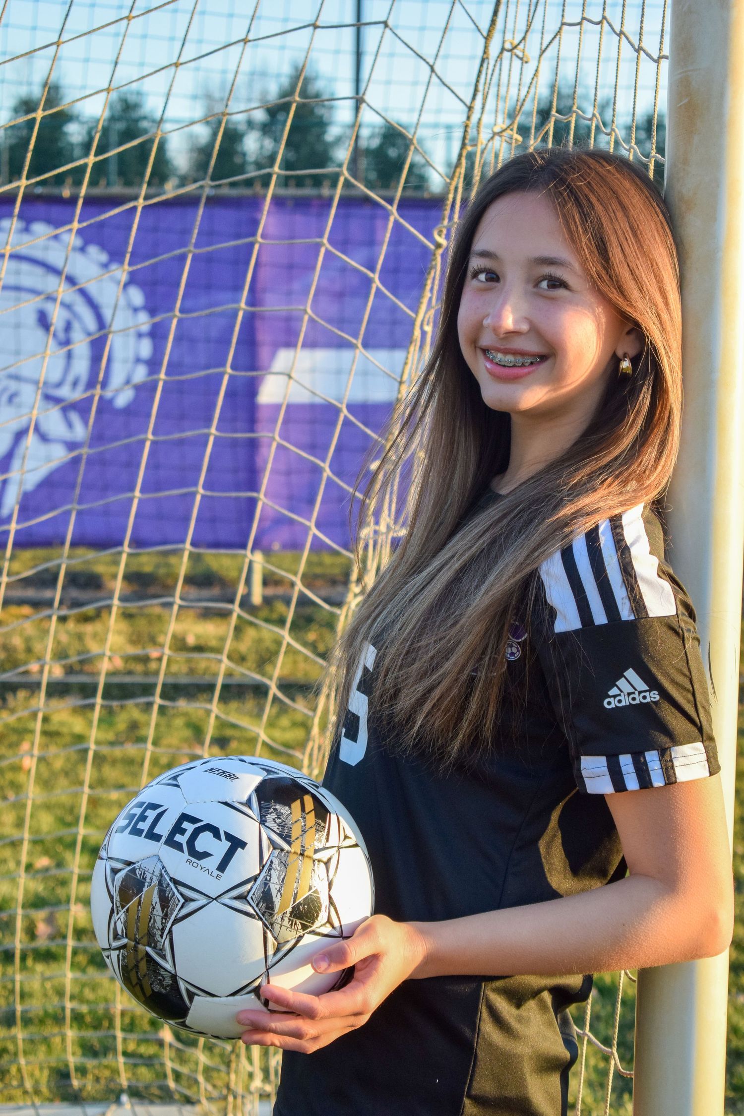 Soccer player in Adidas uniform holding Select ball against goal net and purple background.
