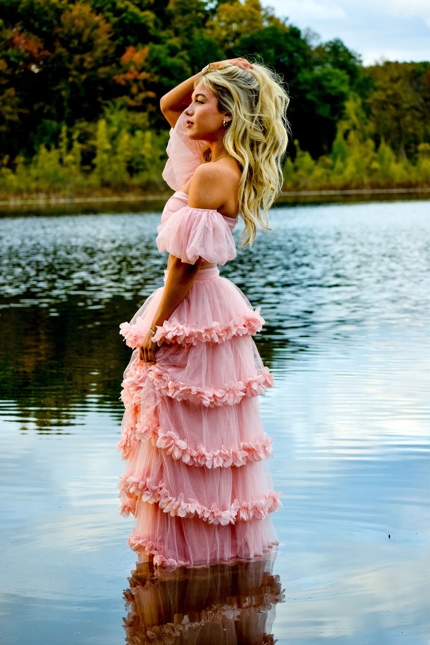 A pink ruffled dress flows gracefully in a lakeside photoshoot with autumn trees reflected in the calm water.