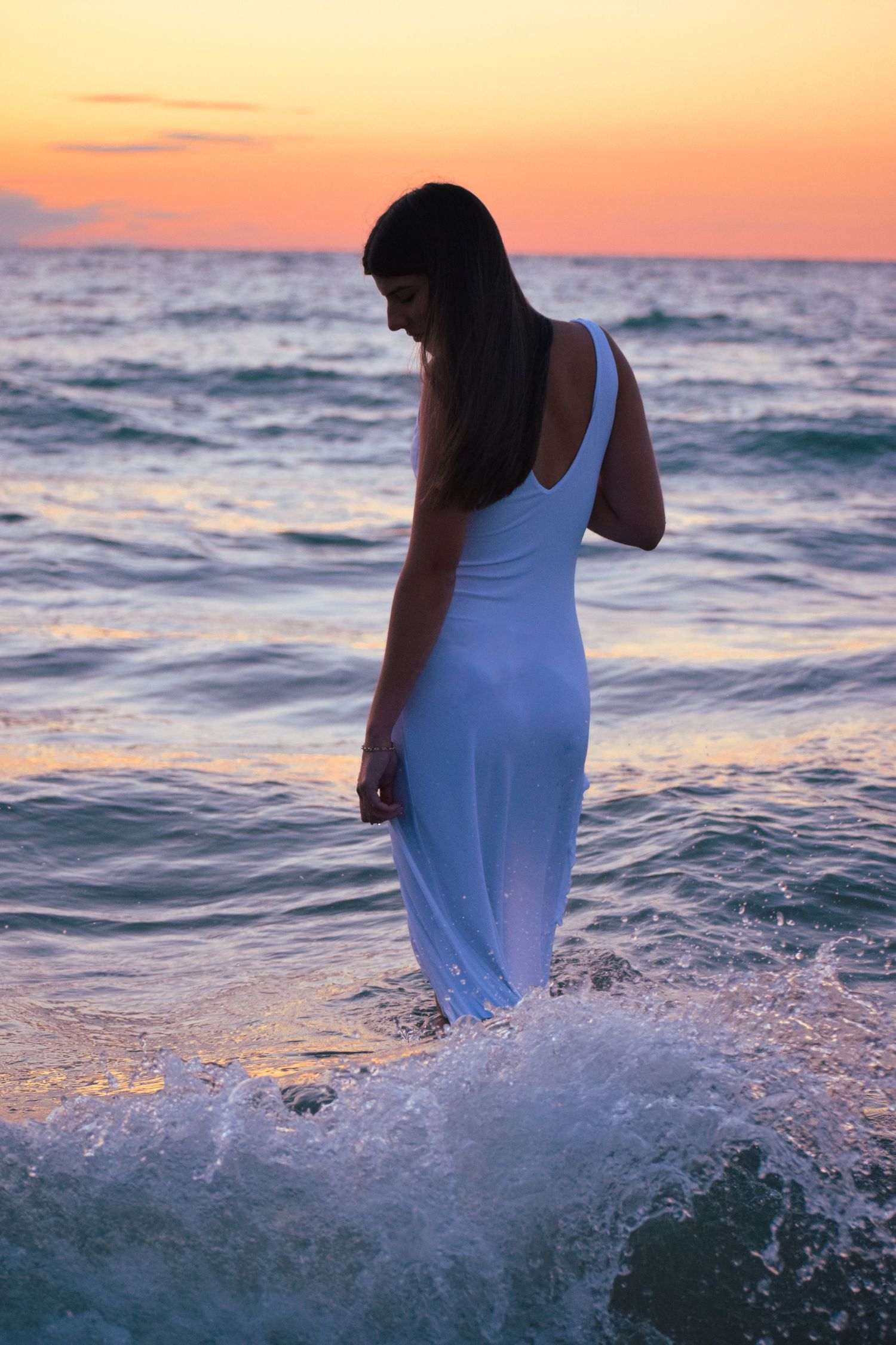 Ocean waves splash around figure in white dress during golden hour photoshoot.