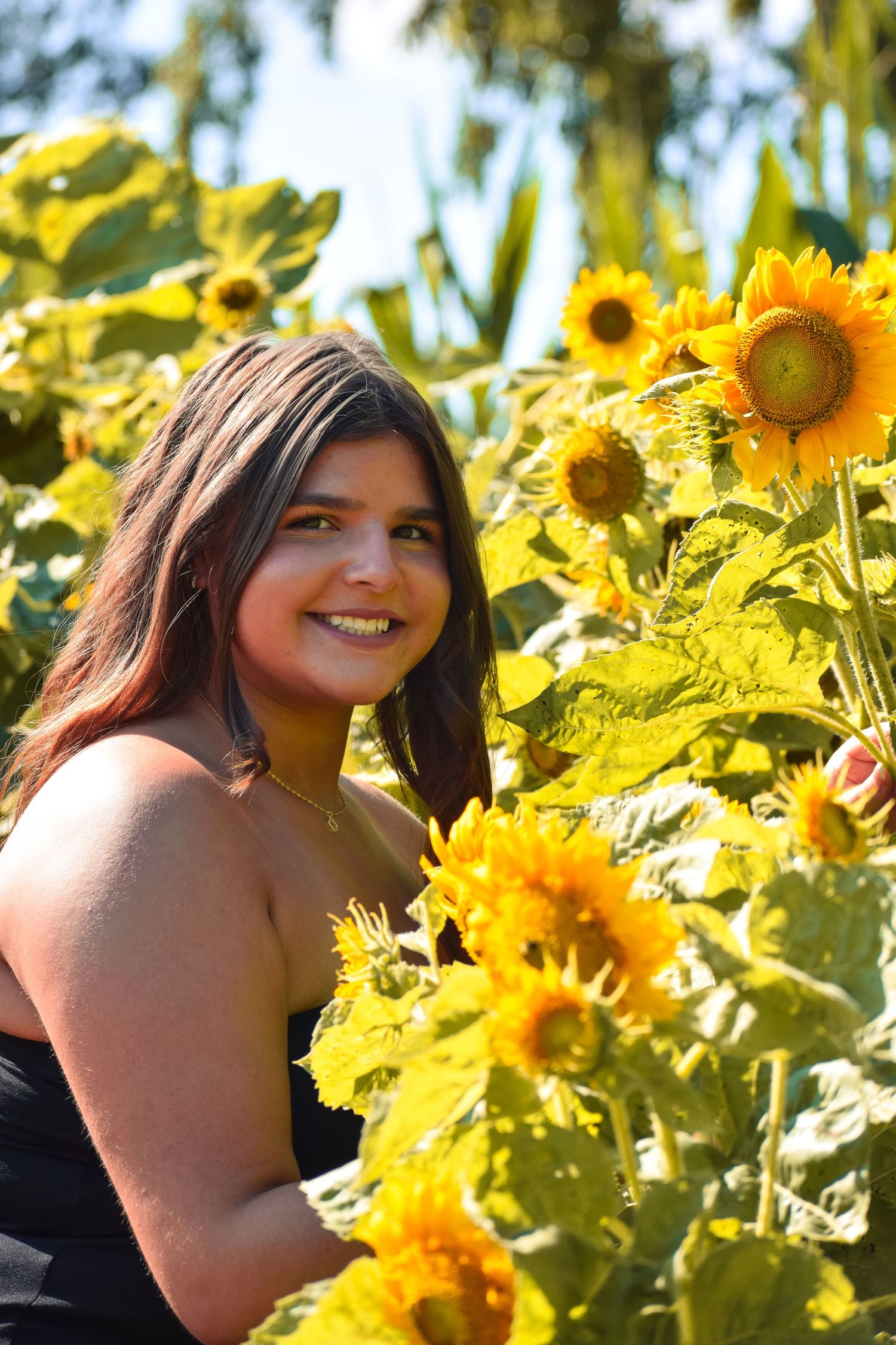 Woman in black top poses among bright yellow sunflowers in sunny garden.