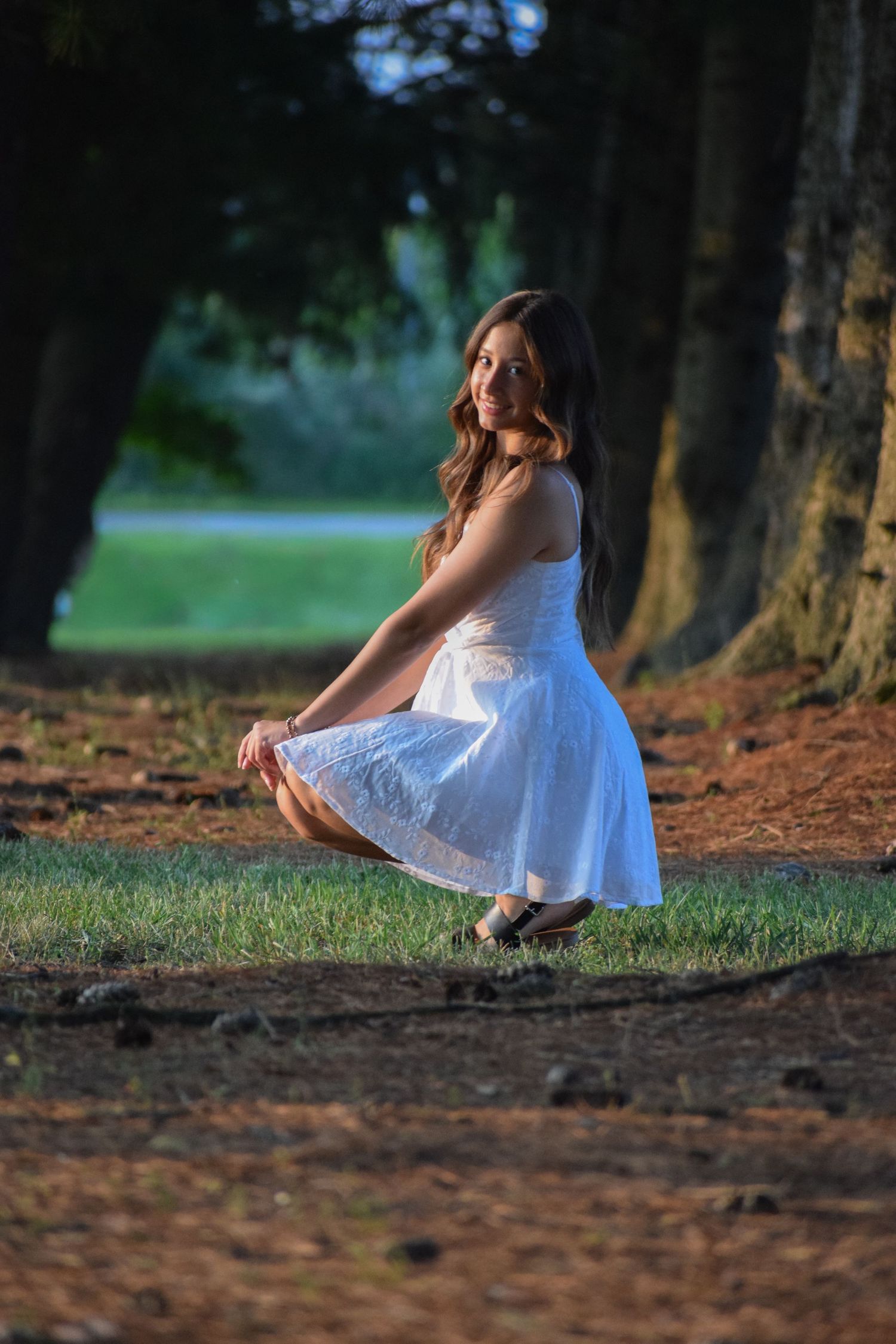 Person in white dress sitting on pine needle covered ground in forest setting.