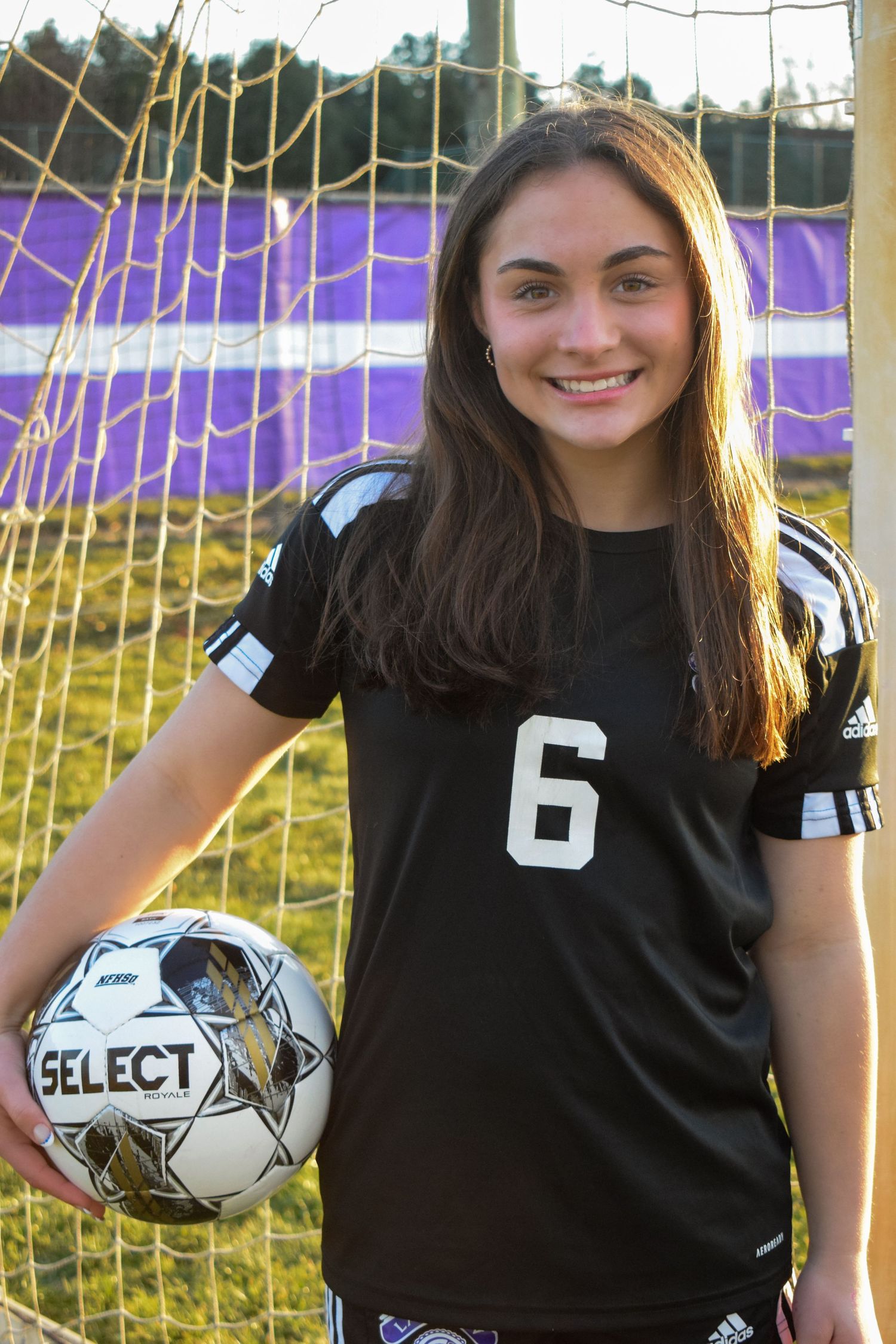 Soccer player in black uniform posing with Select brand ball near purple goal netting.