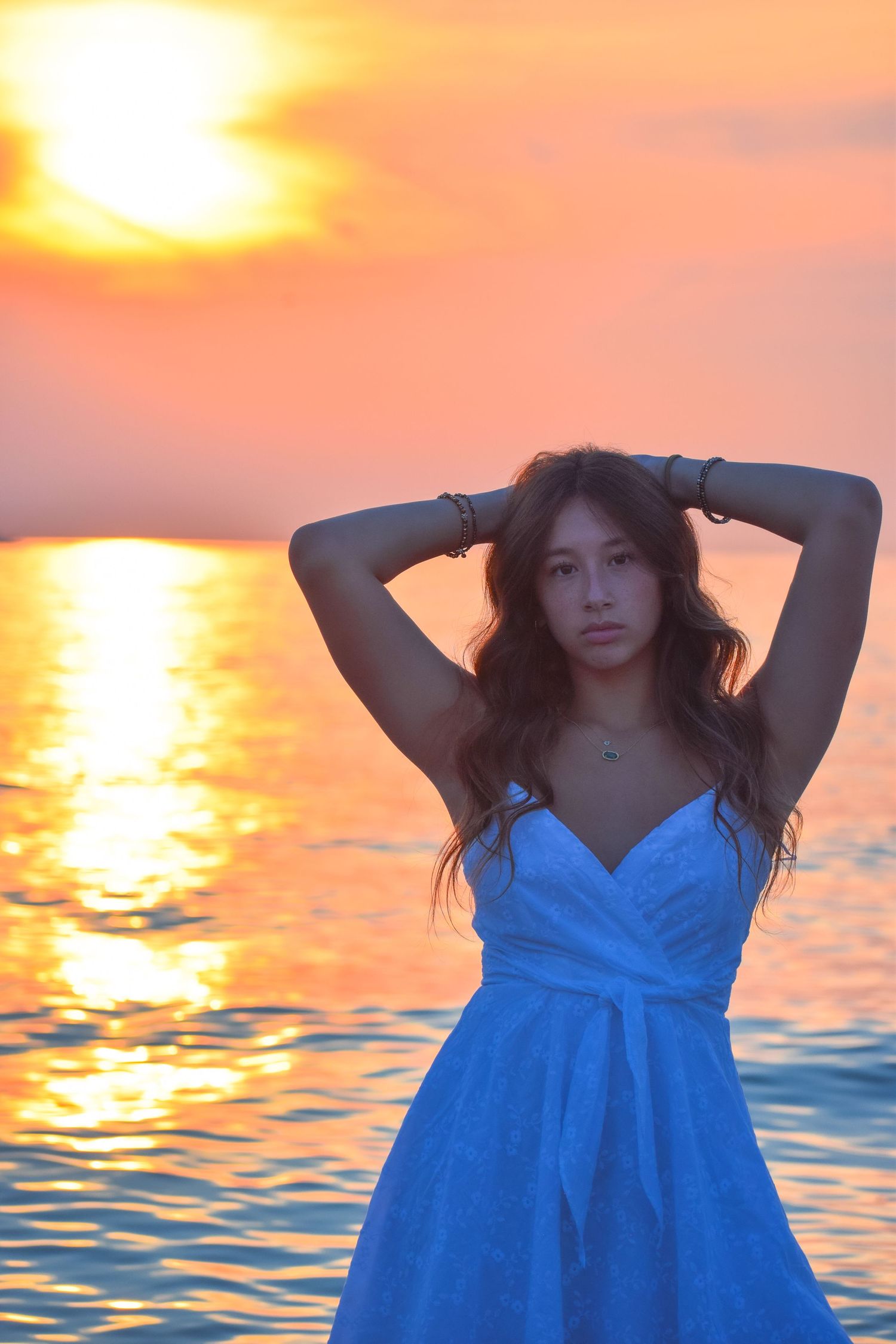 Woman in blue dress poses against dramatic orange sunset over water.