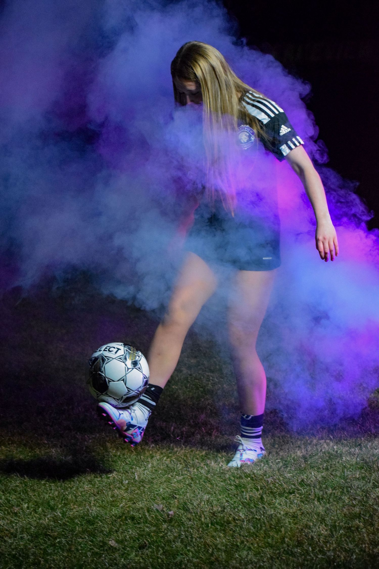 Soccer player kicking a ball in purple smoke on a grass field at night.