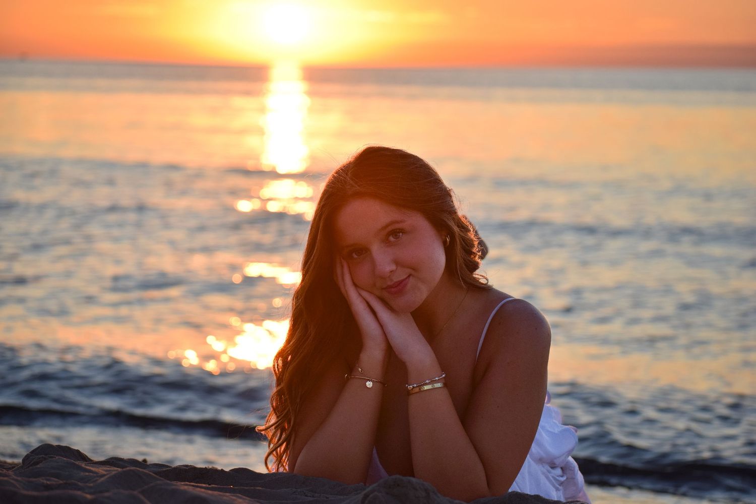 Silhouetted figure relaxing on beach at sunset with orange sky reflecting on calm ocean waters.