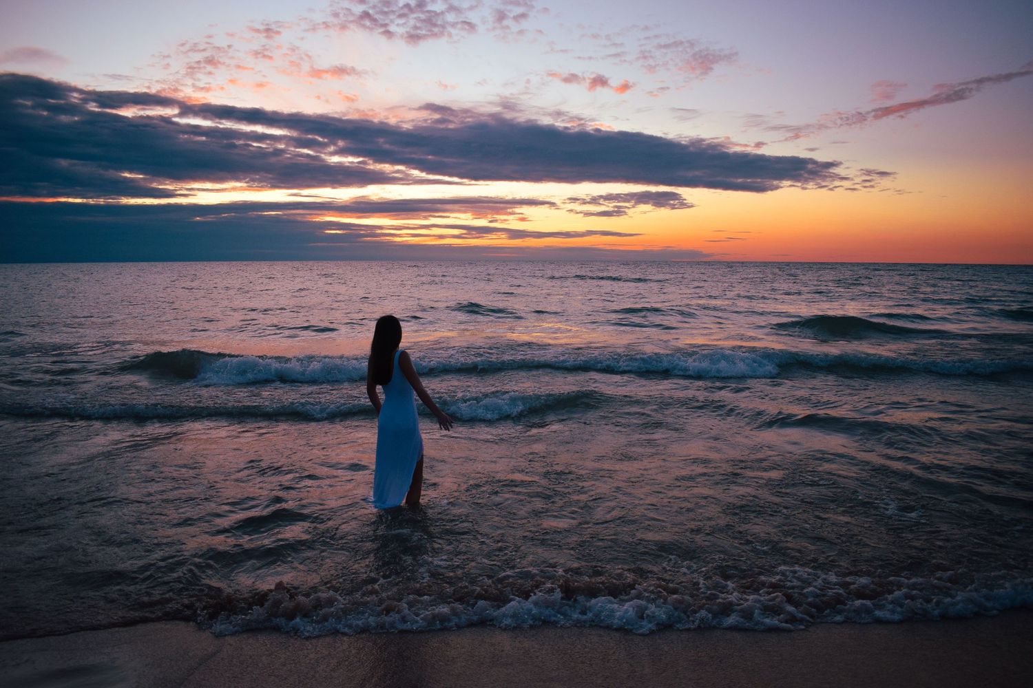 Silhouette stands against dramatic orange and purple sunset over ocean waves.