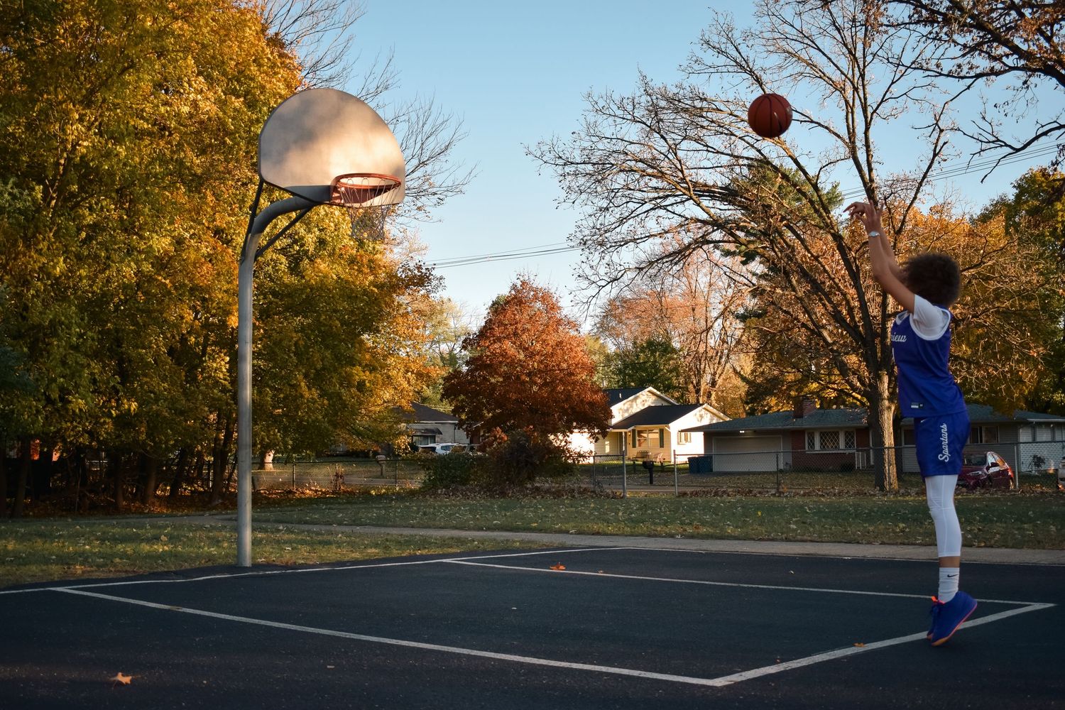 Basketball players practicing on an outdoor court during autumn with golden trees in the background.