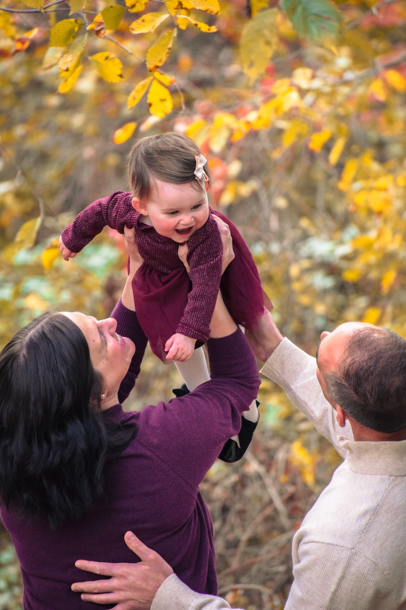 Family playfully lifts child in the air against autumn foliage backdrop in burgundy sweater.